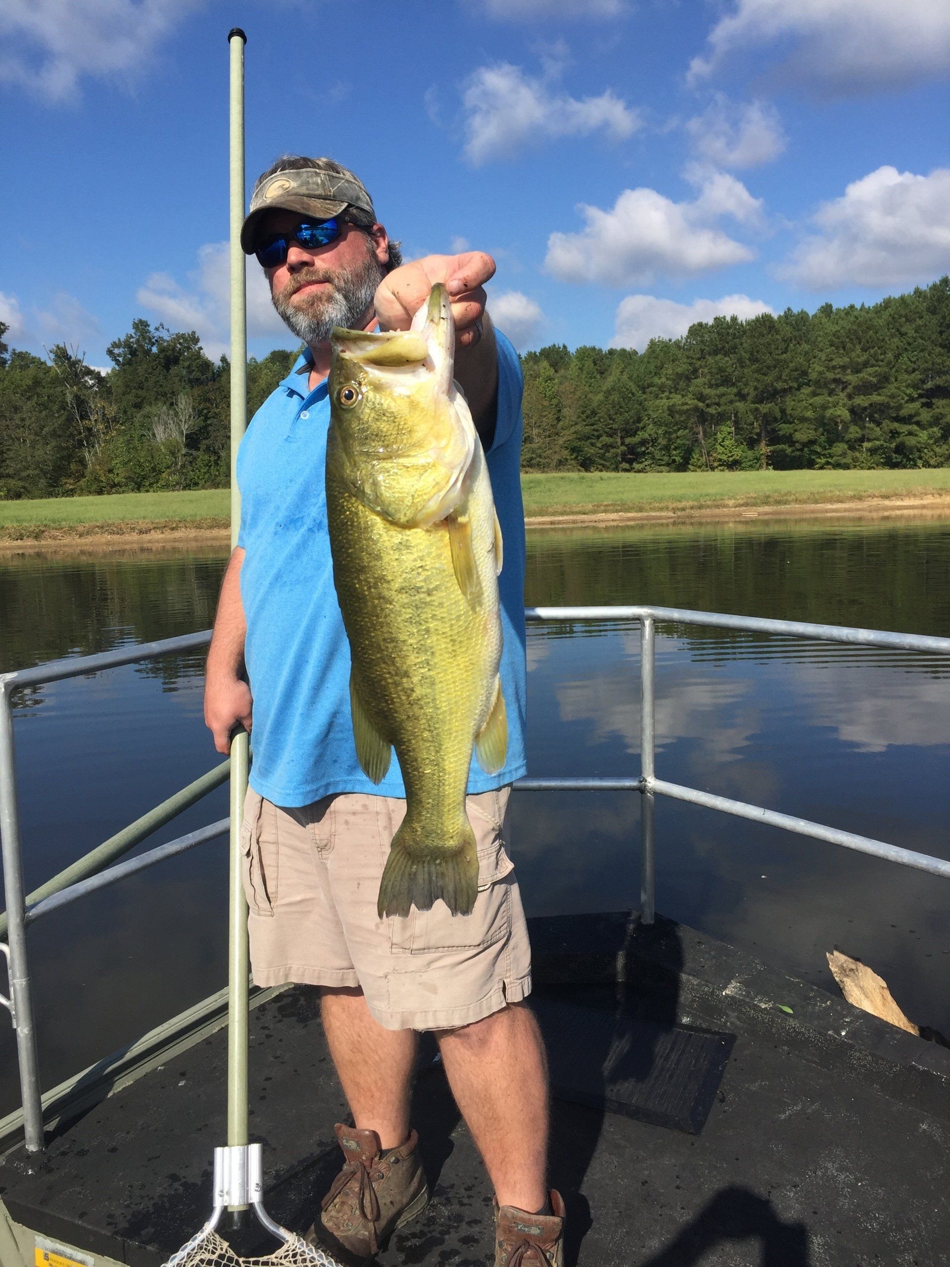 Man holding a large bass he caught on a boat, near a lake with trees.