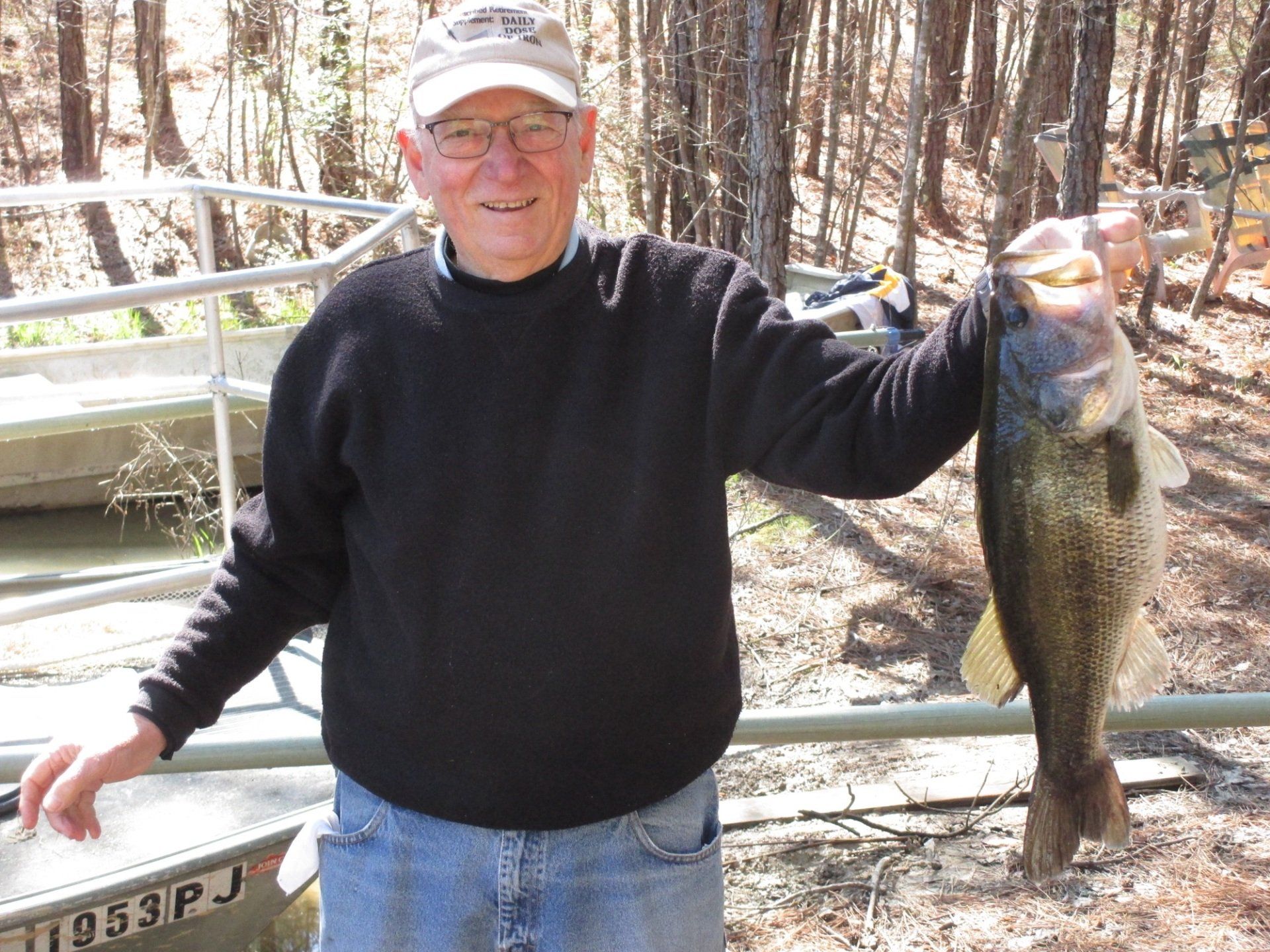 Man smiles, holding large fish. Outdoors, near a boat. Black sweater, hat. Trees in the background.
