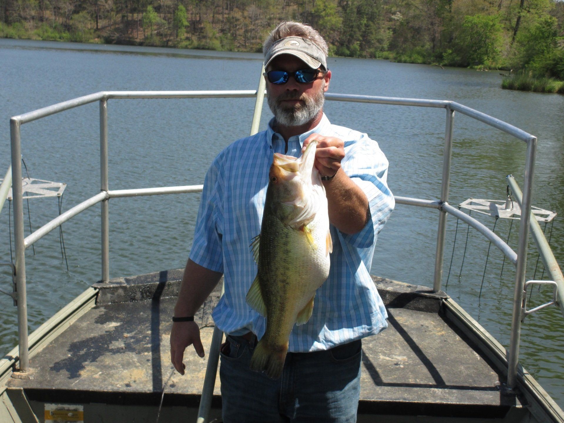 Man on a boat holding a large fish he caught, with a lake in the background.