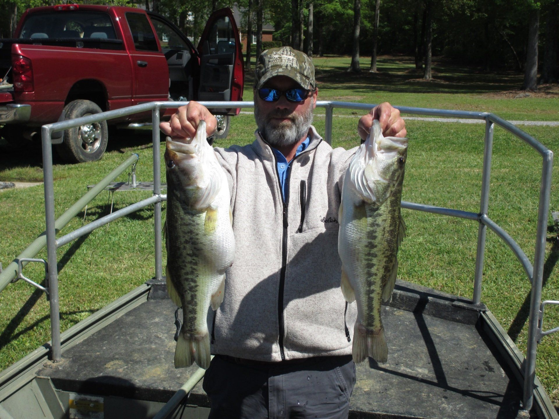 Man holding two large bass fish on a boat, red truck in the background.