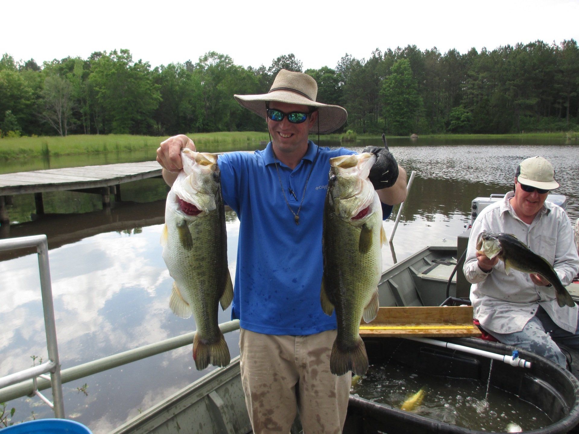 Man holding two large bass, another person in a boat holding one, at a lake.