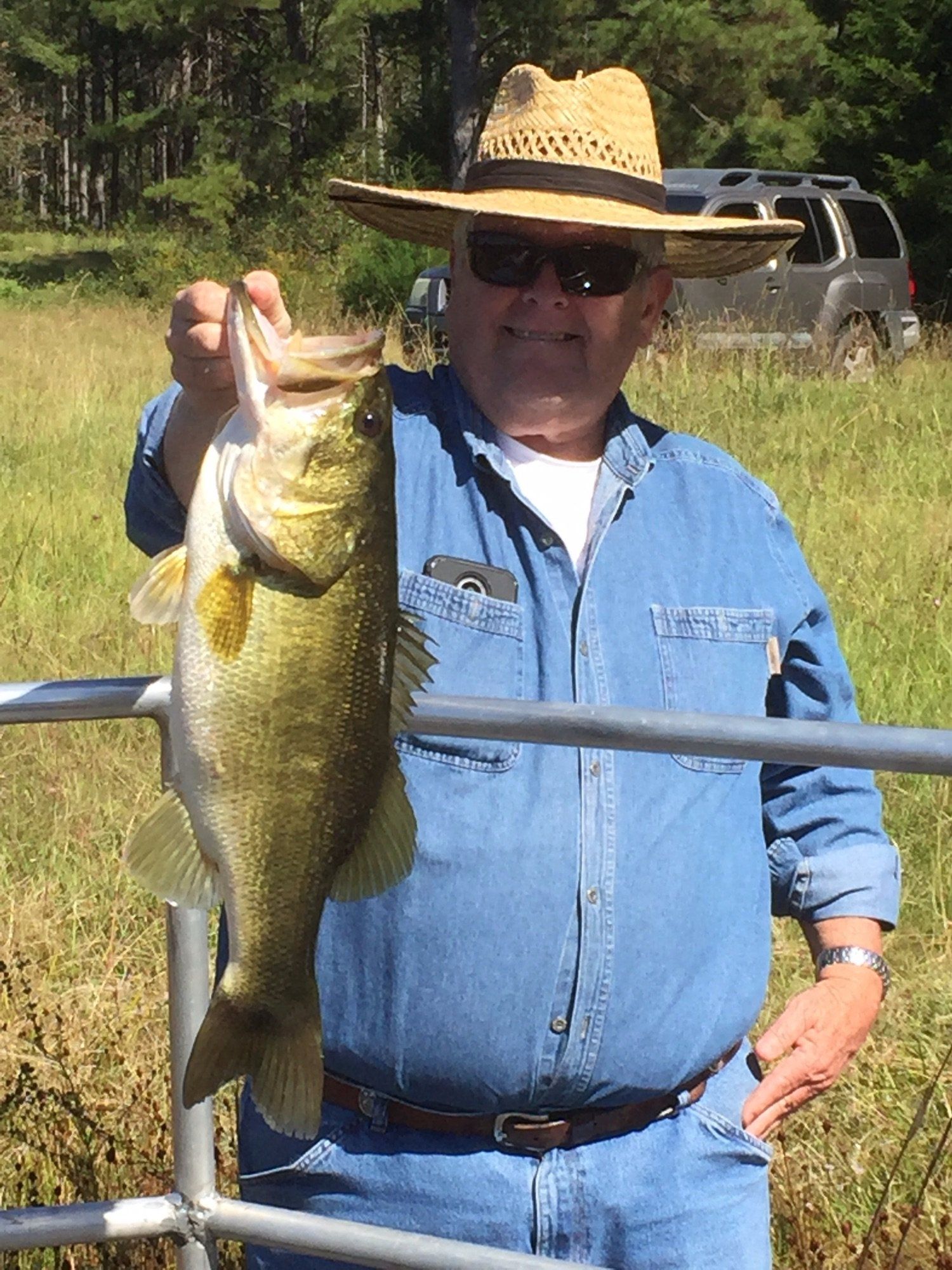 Man in straw hat holds up a large bass, smiling. Outdoors near a metal rail.