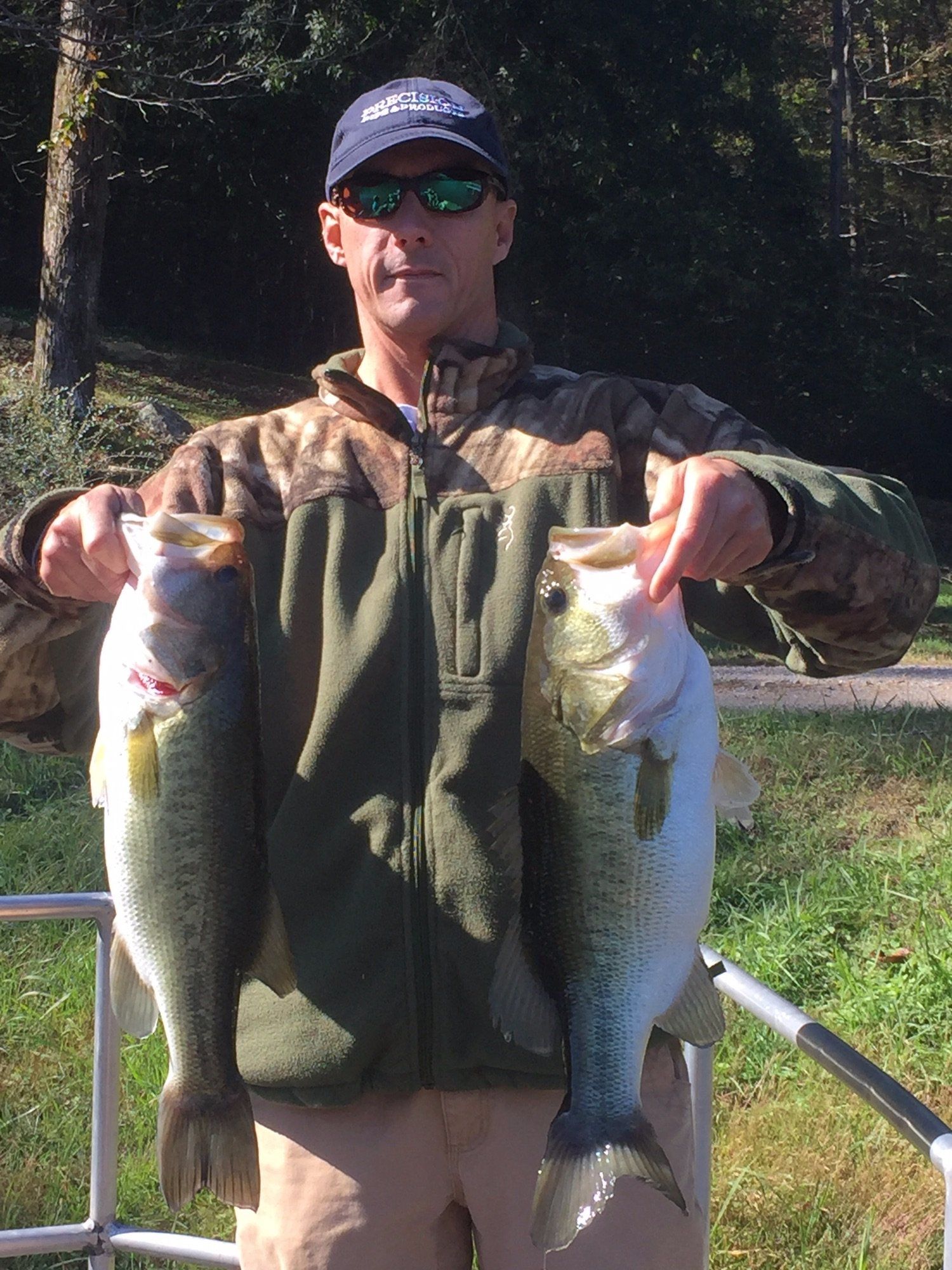 Man holding two large bass; outdoors, pointing at the fish, wearing camouflage jacket and cap.