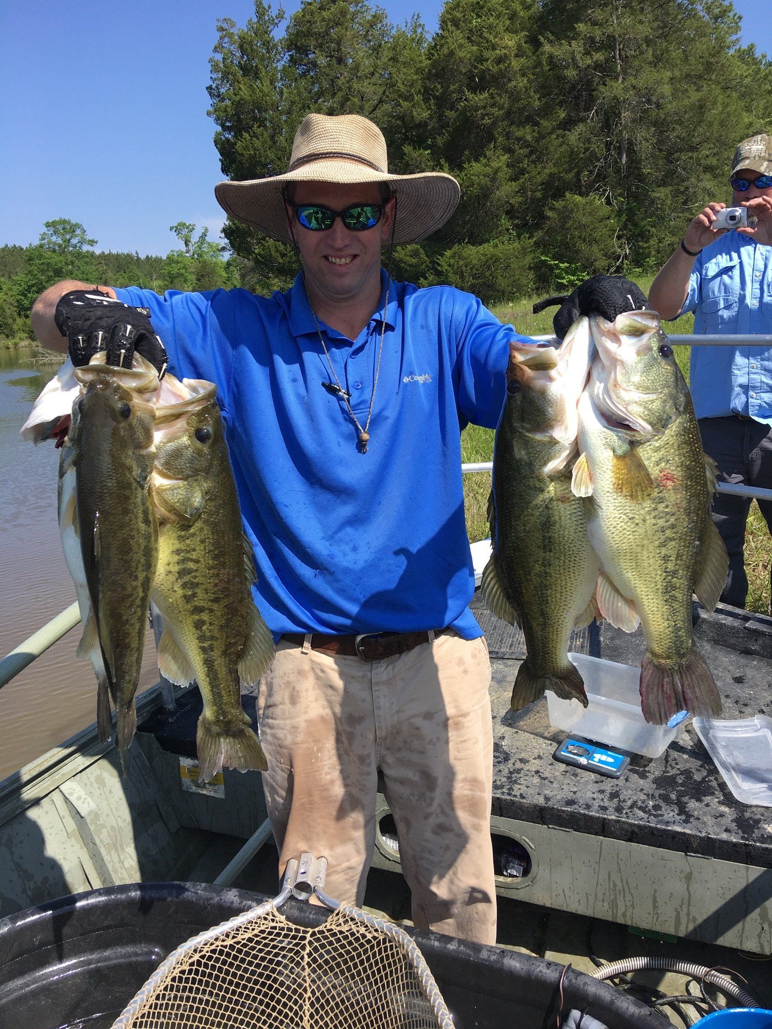 Man holding four large bass on a boat, wearing a blue shirt, khaki pants, and a straw hat, sunny day.