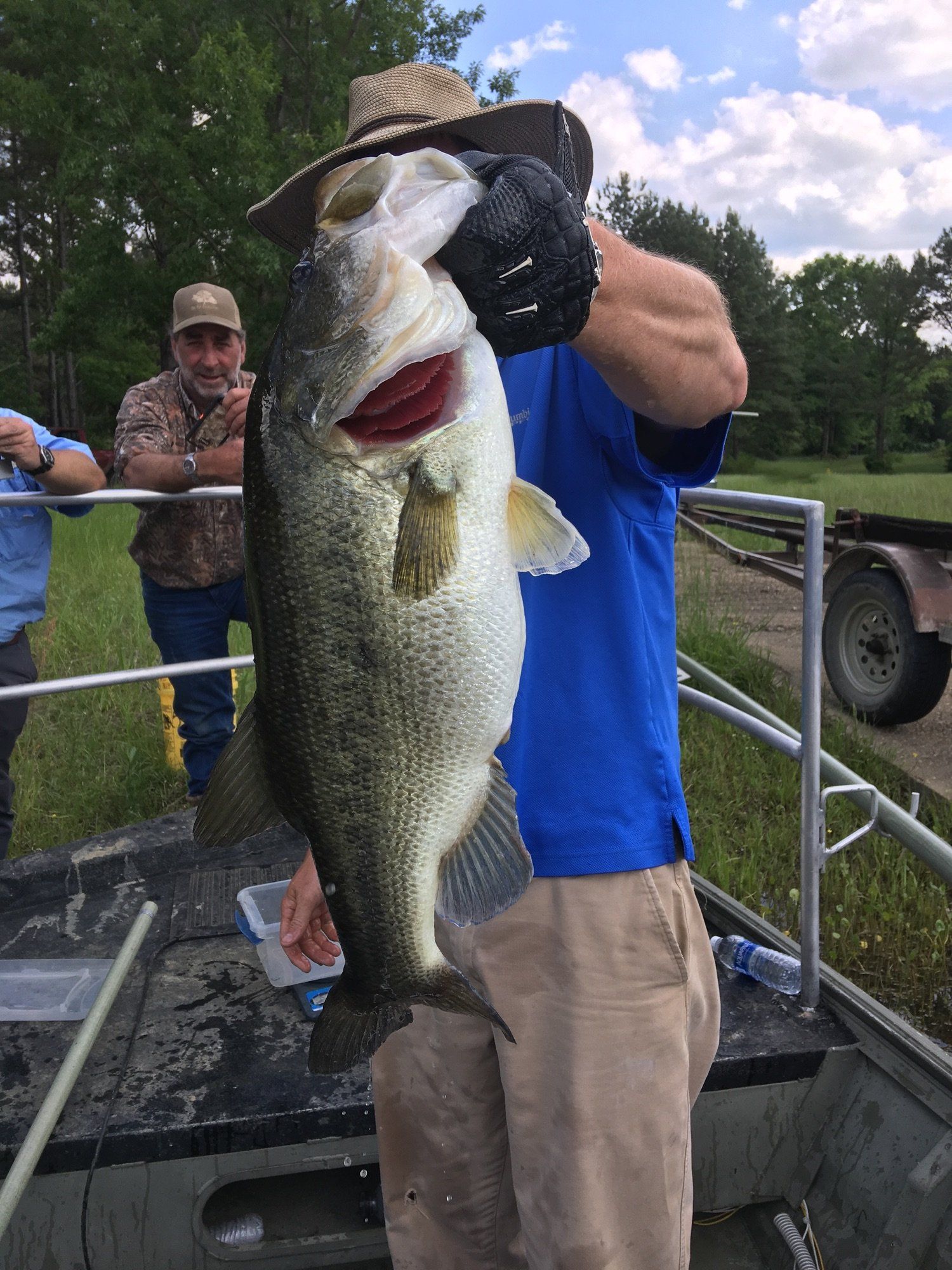 Man holding a large bass fish on a boat, other men in the background, outdoors.