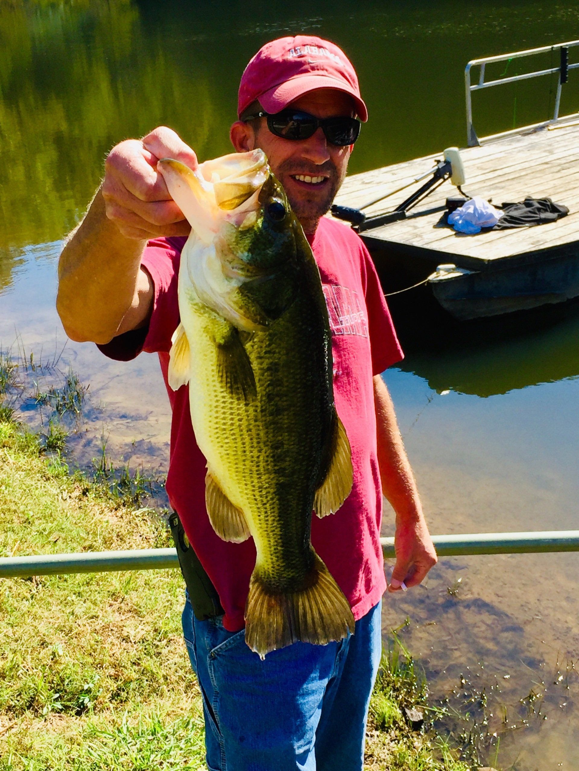 Man holding up a large bass, smiling. Red cap, t-shirt, blue jeans, lake background.