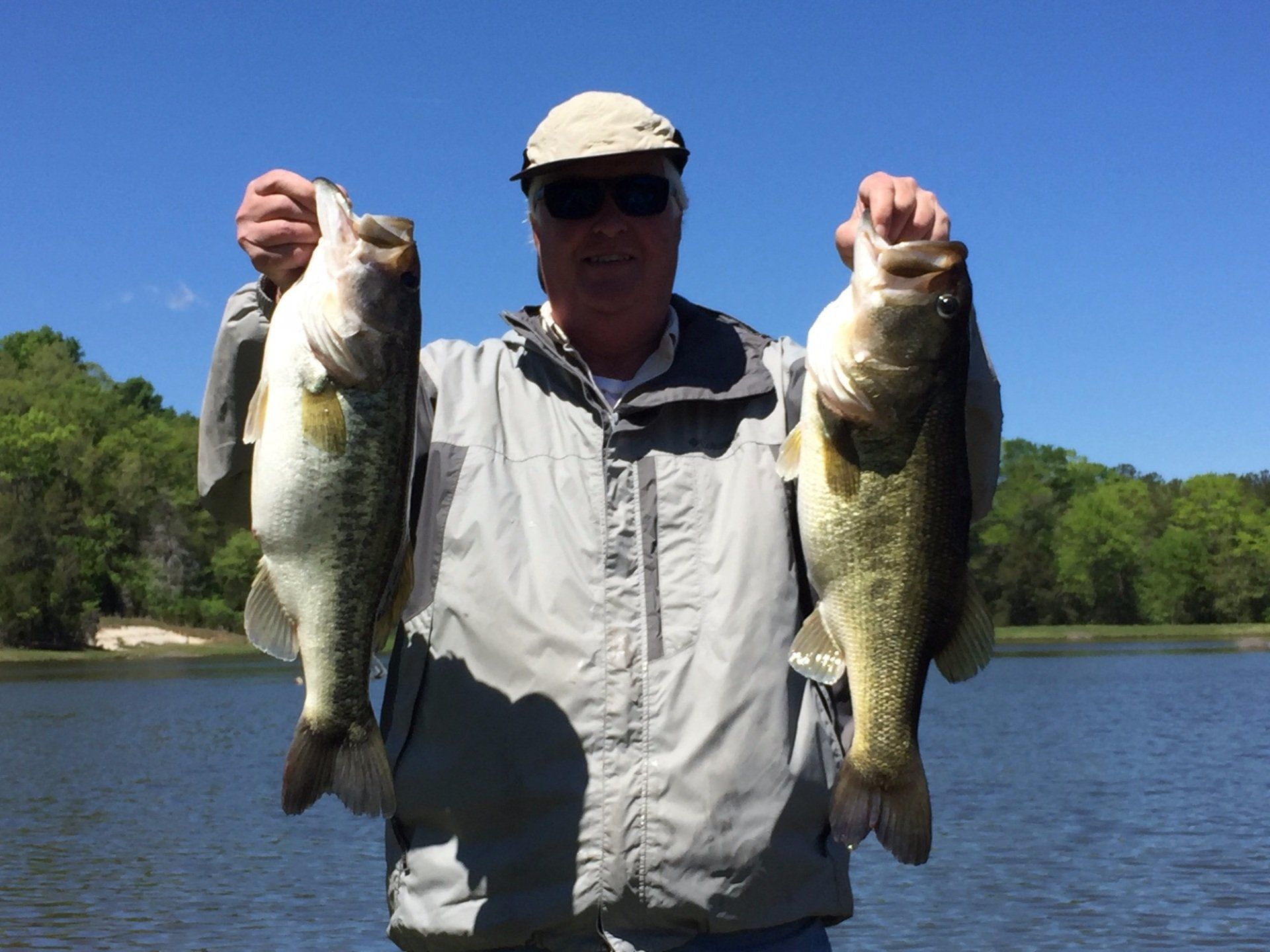 Man holding up two large bass he caught, standing in a lake with a blue sky and trees.