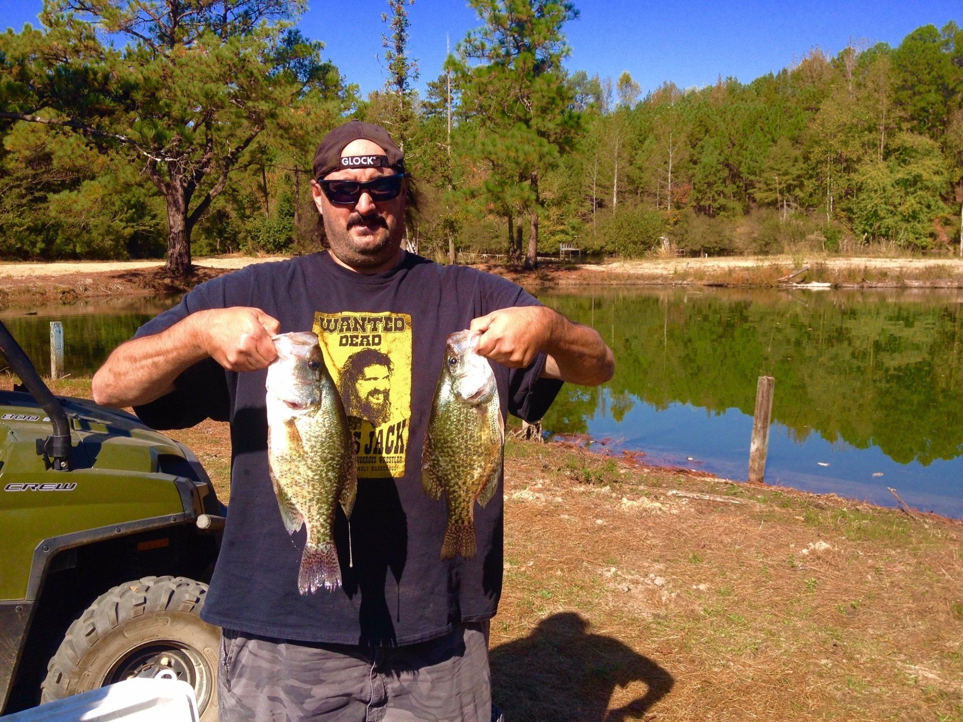 Man holding two fish by a pond, wearing sunglasses, with a four-wheeler in the foreground.