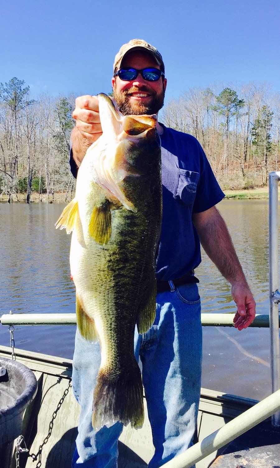 Man holding a large largemouth bass on a boat, smiling. Outdoors near water and trees, sunny day.