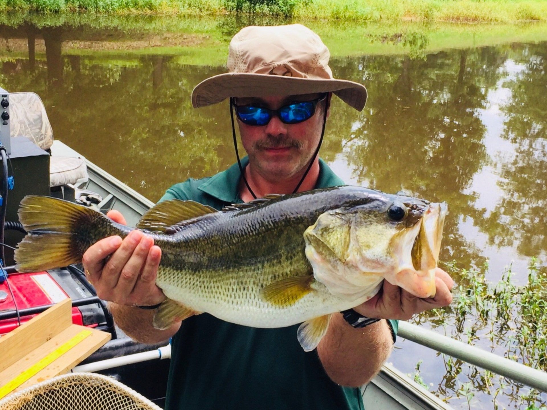 Man in hat holding a large bass fish with an open mouth on a boat.