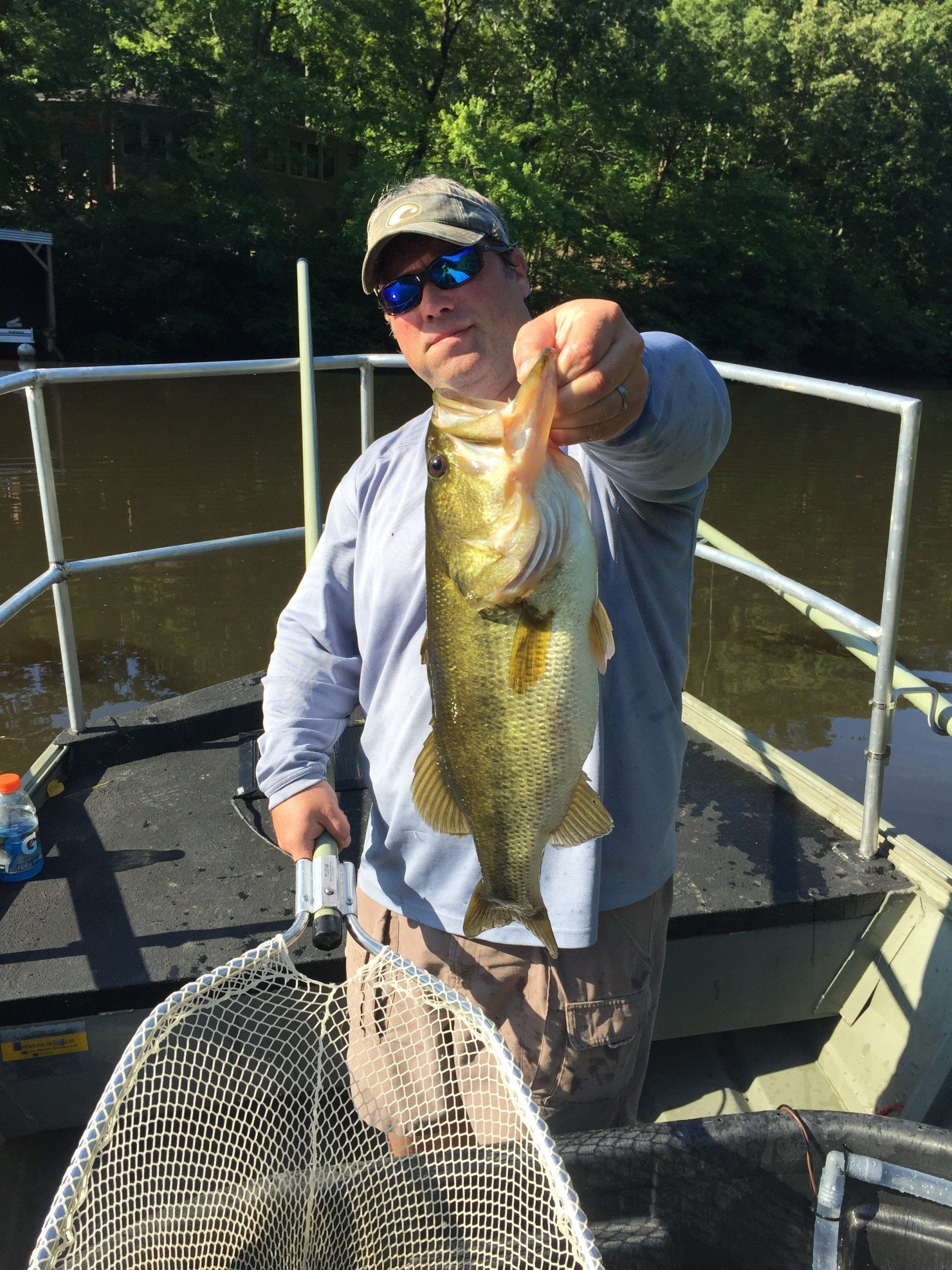 Man in a fishing boat holding up a large bass; green water and trees in background.