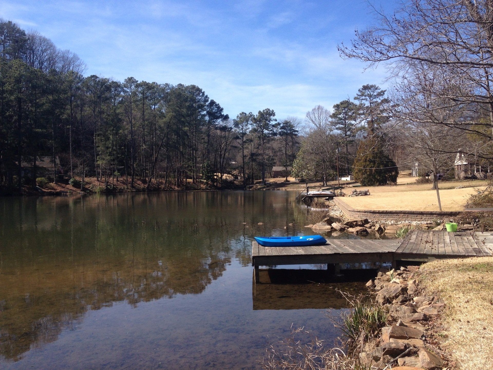 A lake with a dock, trees, and blue sky. A kayak sits on the dock, and houses line the far shore.