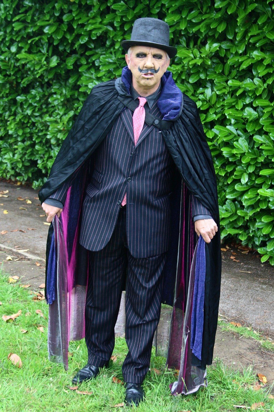 A man in a suit and cape is standing in the grass holding umbrellas.