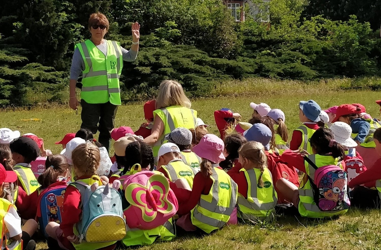 A woman in a green vest is talking to a group of children sitting on the grass.