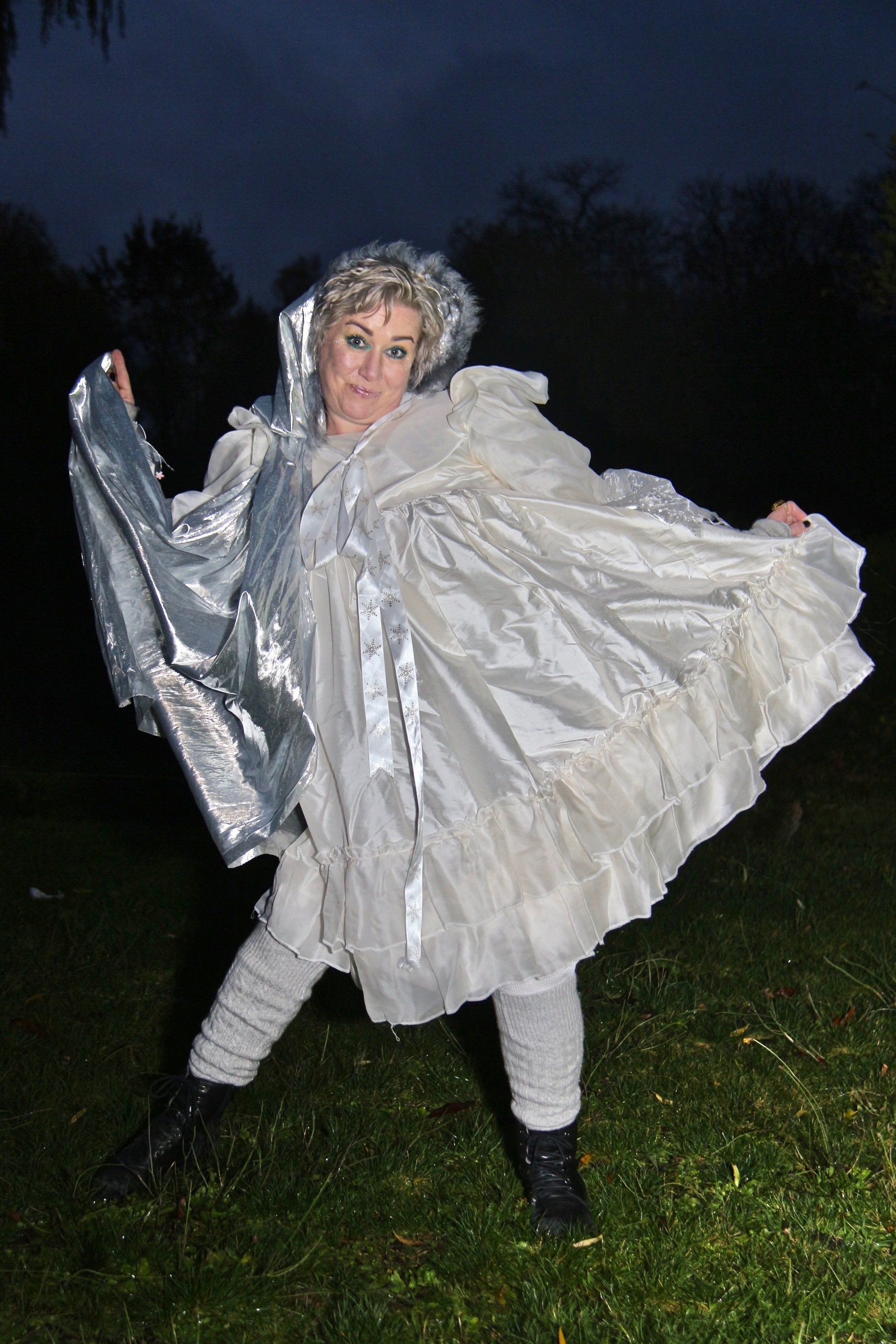 A woman in a white dress and silver cape is standing in the grass.