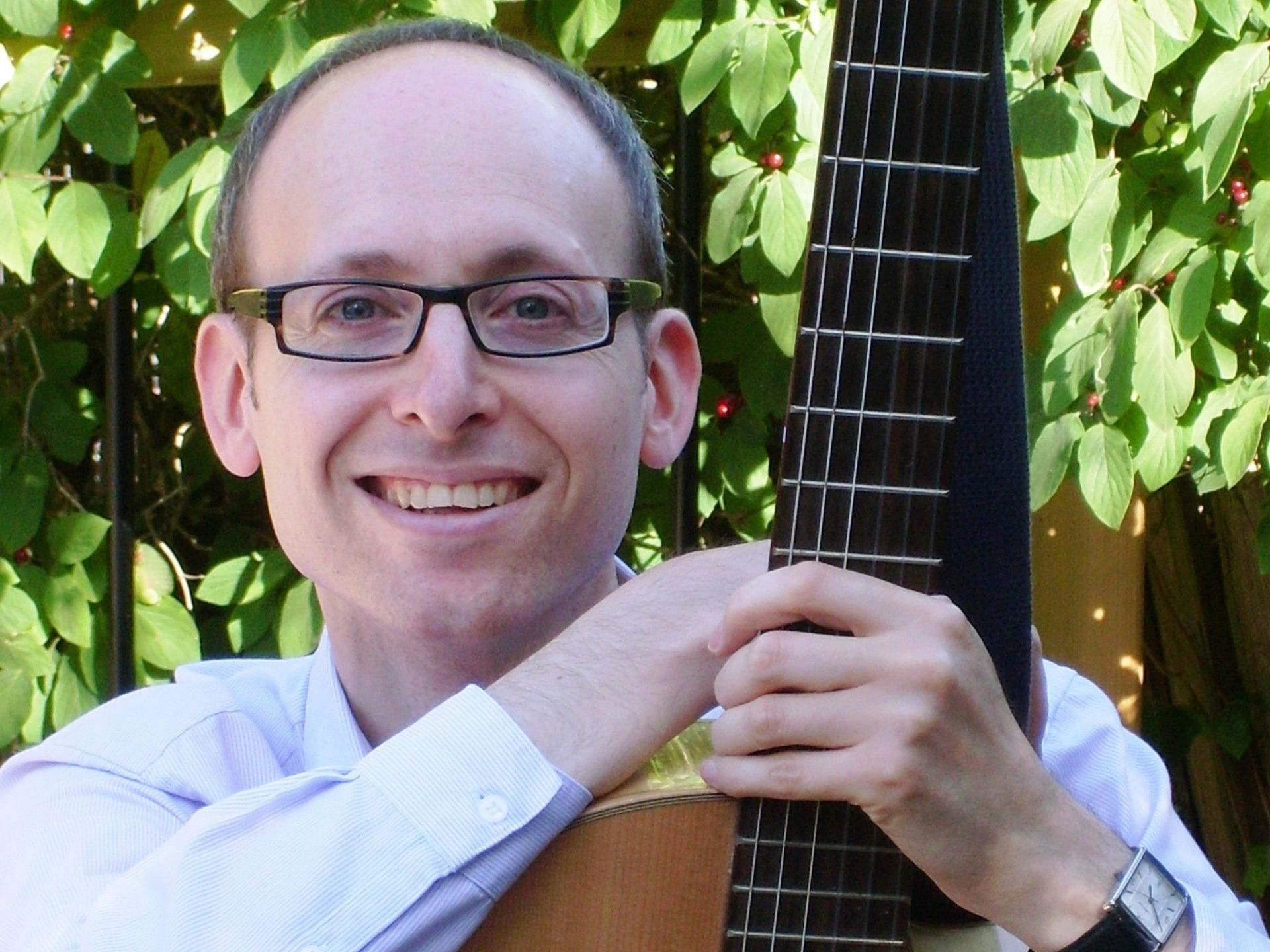 A man wearing glasses is holding a guitar and smiling