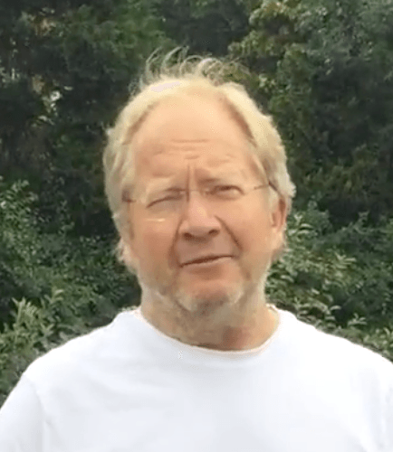A man with glasses and a beard is wearing a white shirt and standing in front of trees.