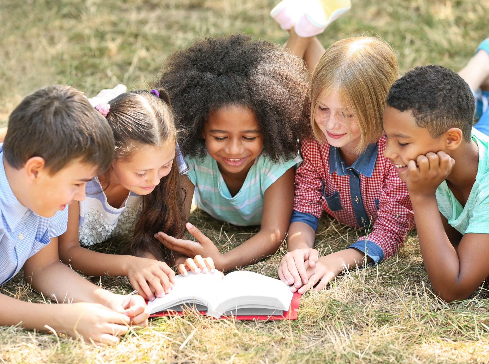 A group of children are laying on the grass reading a book.
