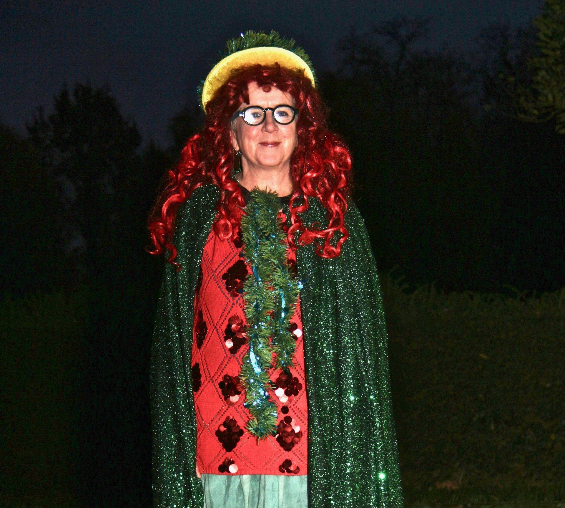 A woman with red hair and glasses is wearing a green cape and a red sweater