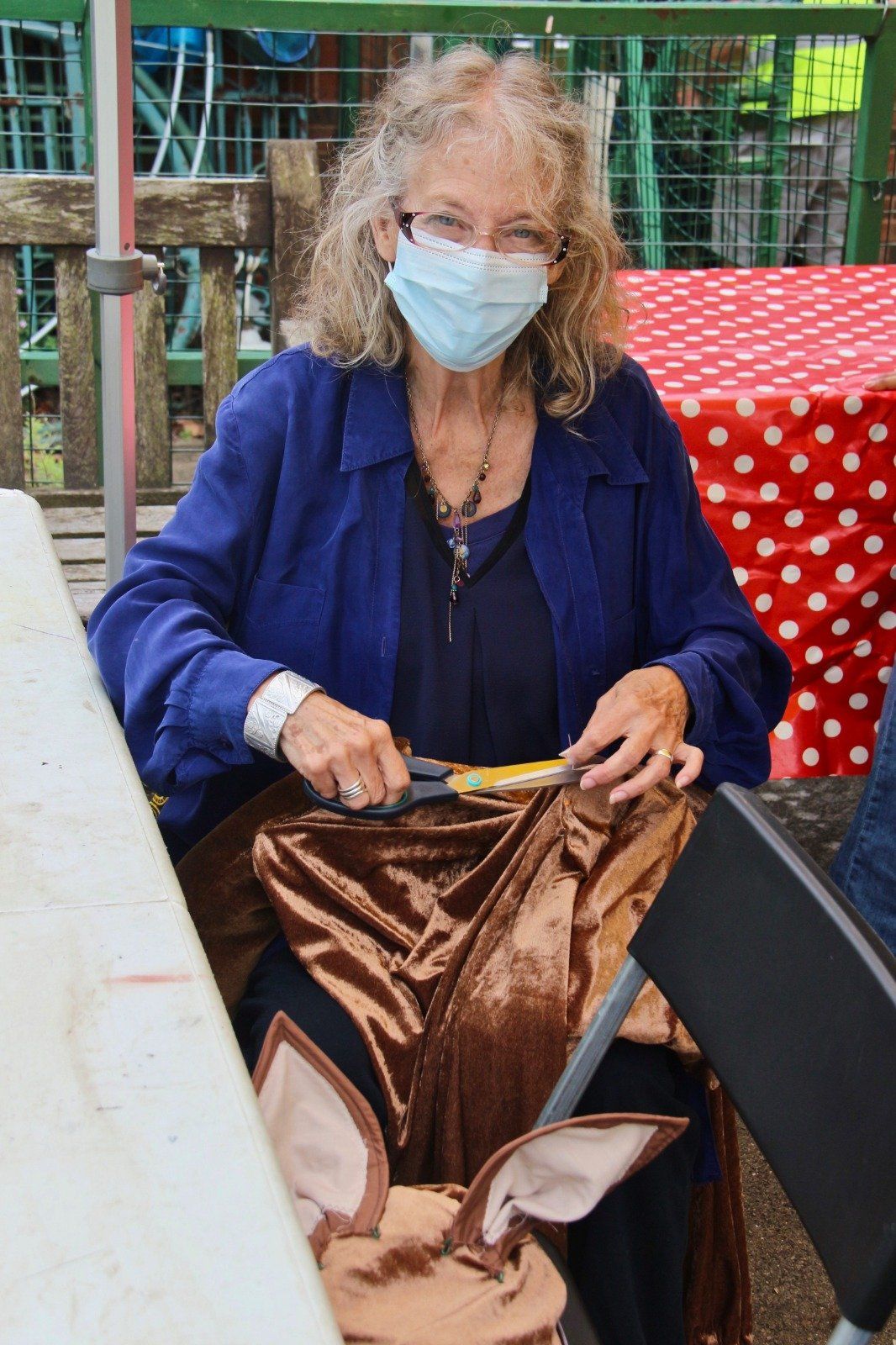A woman wearing a mask is sitting at a table cutting a piece of fabric.