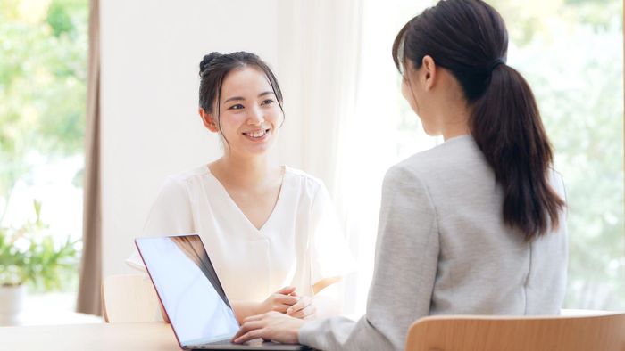 Two women are sitting at a table with a laptop and talking to each other.