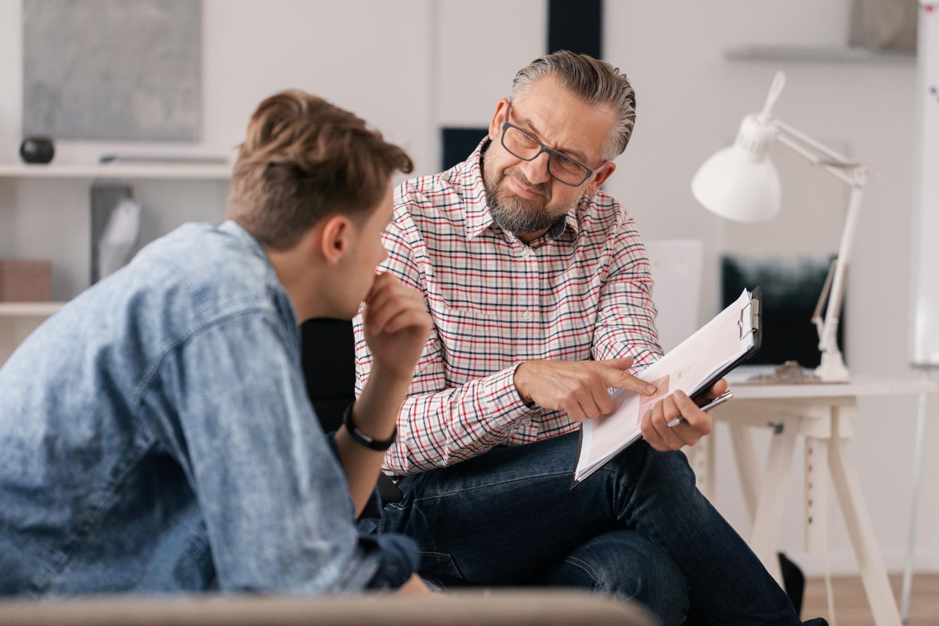 A man is sitting on a couch talking to a young man while holding a clipboard.