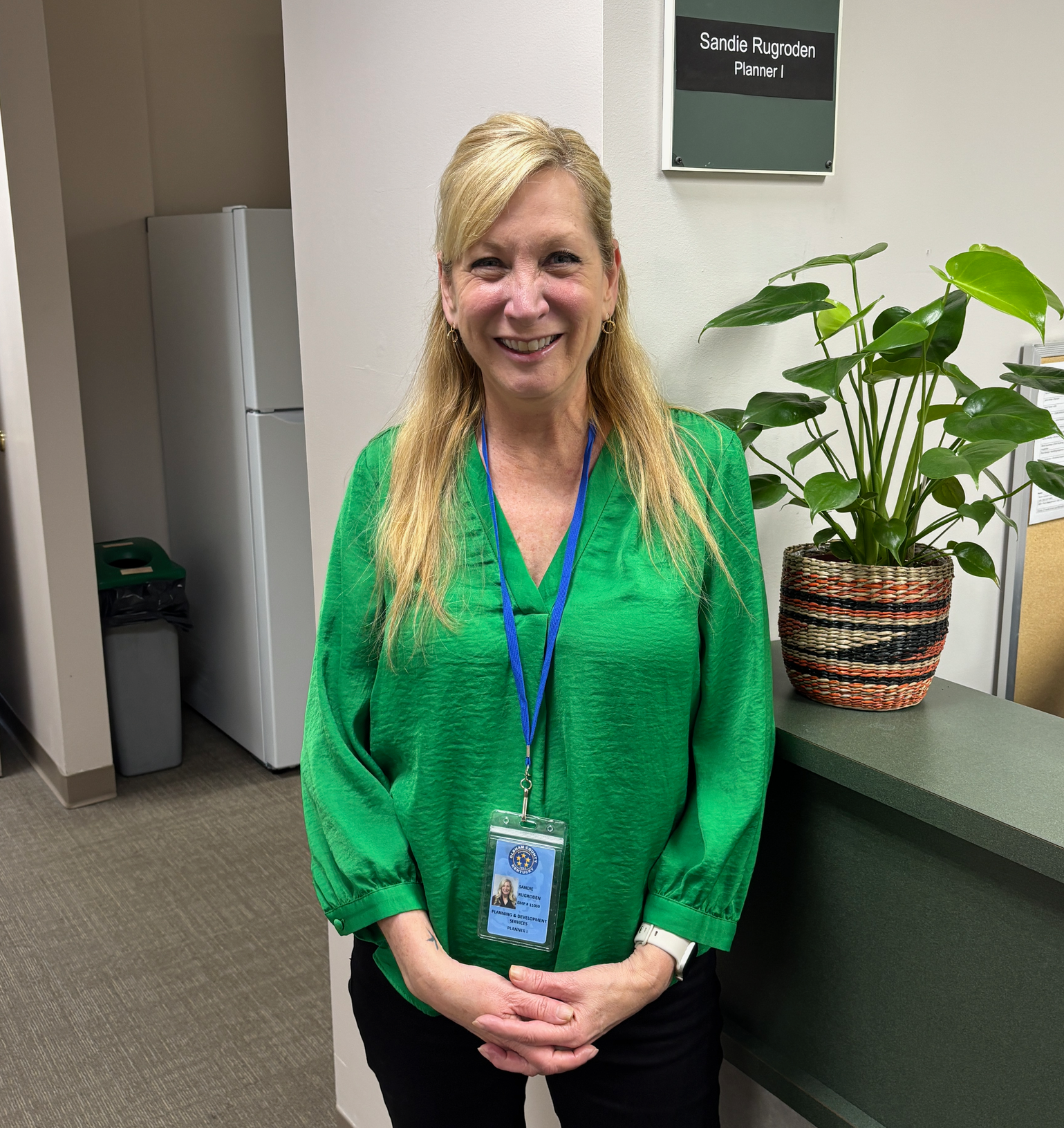 A woman in a green shirt is standing next to a potted plant.
