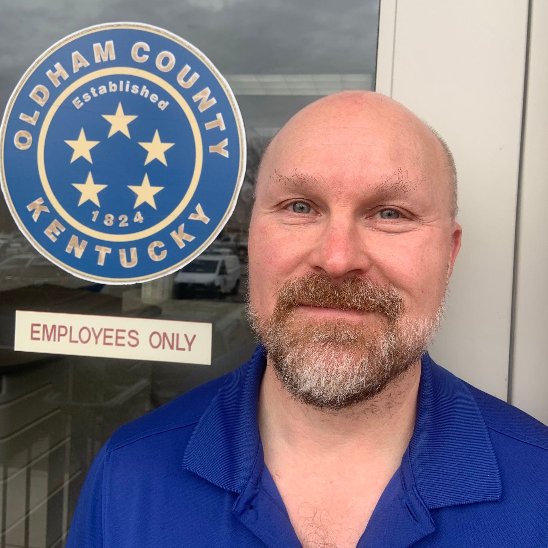 A bald man with a beard is standing in front of an oldham county kentucky logo