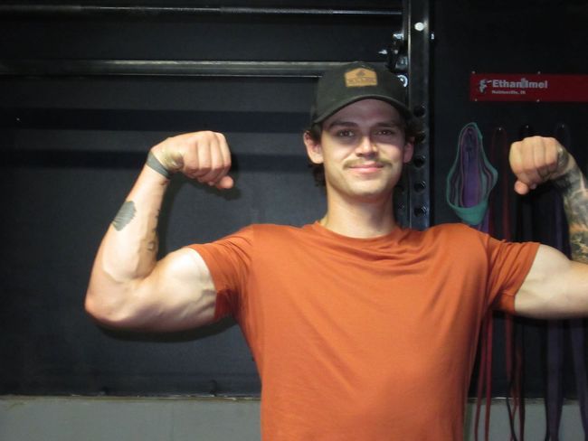 A man in an orange shirt and black hat is flexing his muscles