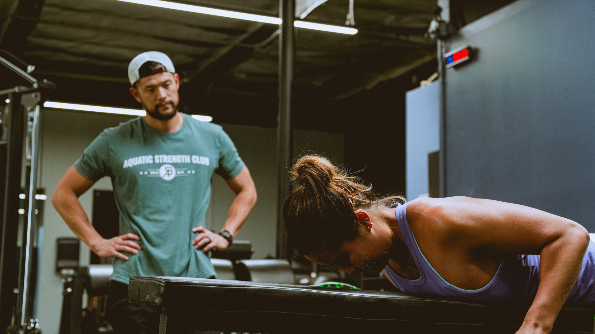 Man in green shirt watches woman in purple tank top doing a bench row in a gym.