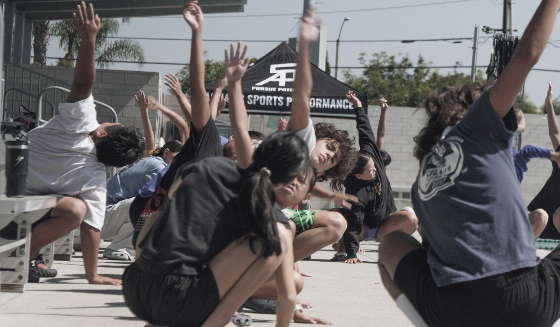 A group stretches outdoors with arms raised, sunny day. Tent in background says 