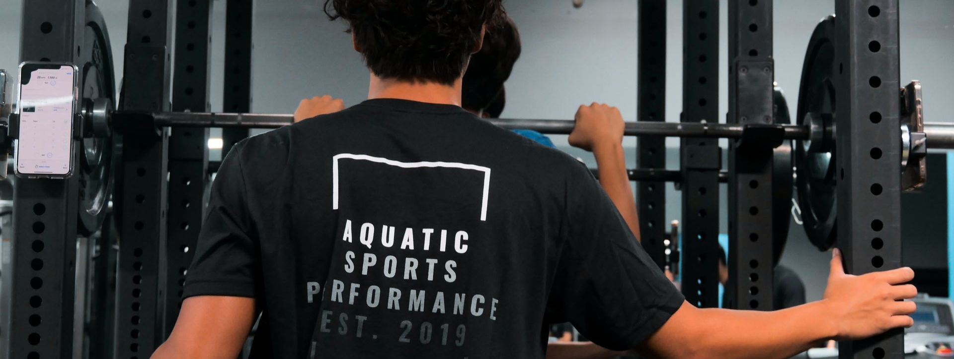 Person in black shirt using a weight rack, holding a barbell. Gym setting.