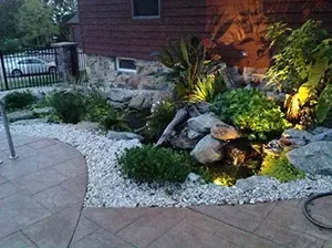 Landscaped garden bed with rocks, plants, and small water feature, illuminated at night.
