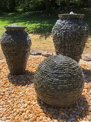 Three stone fountains, two urn-shaped, one round, with water flowing, set on gravel.