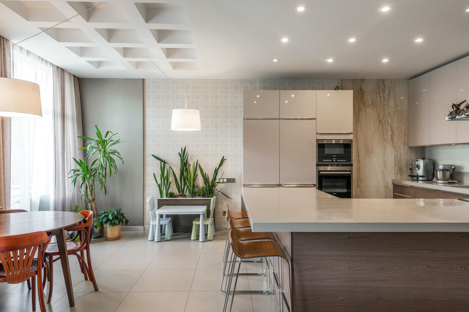 Modern kitchen with island and dining area, light wood and white cabinets, tiled floors, plants.