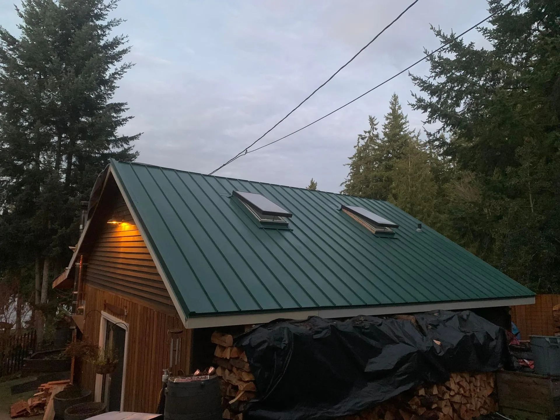 Green metal roof on a small wooden building with two skylights; firewood covered in black tarp in front.