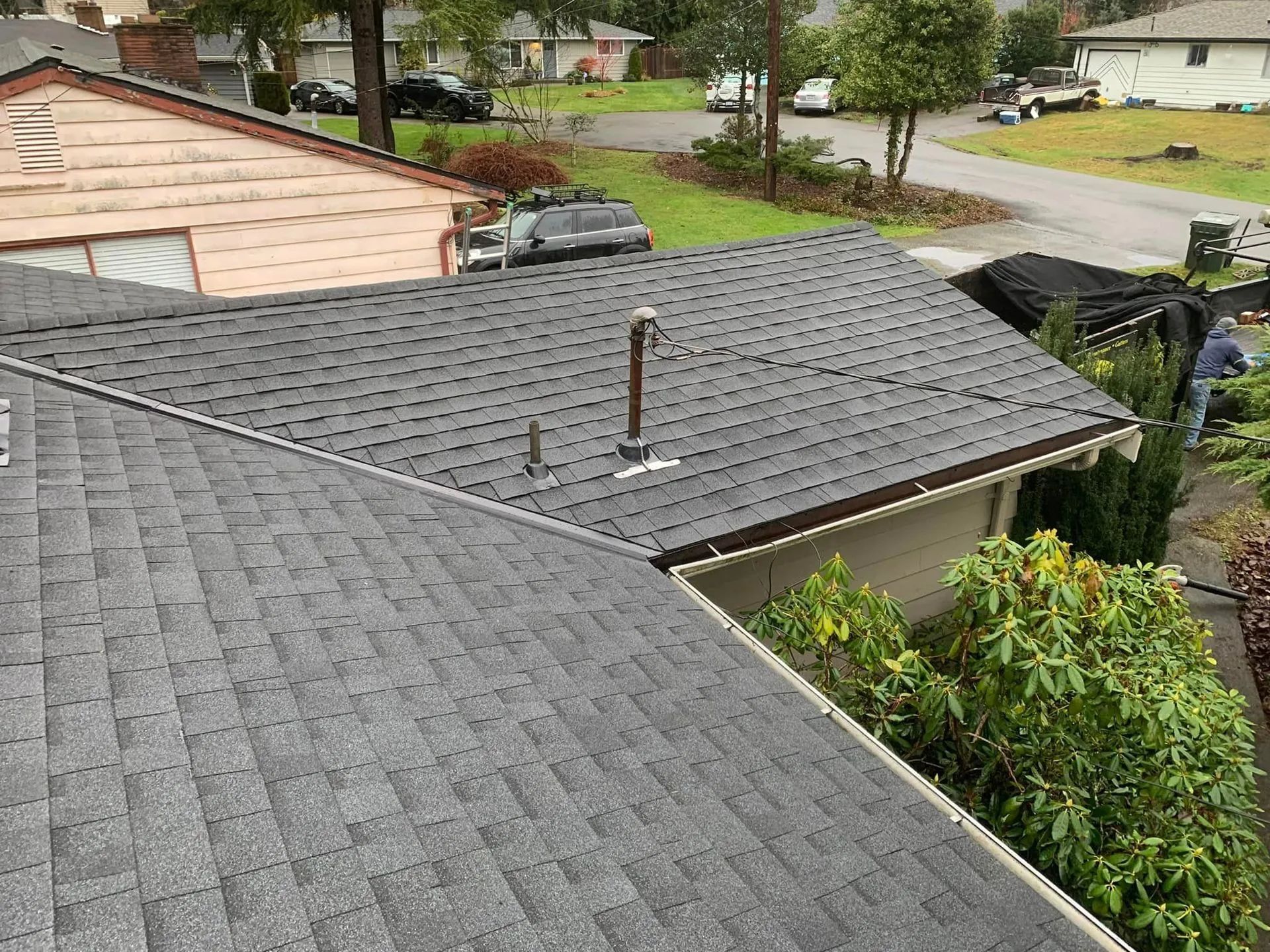 Gray shingle roofs of two houses. Person trimming bushes by the second roof, overlooking a street and other houses.