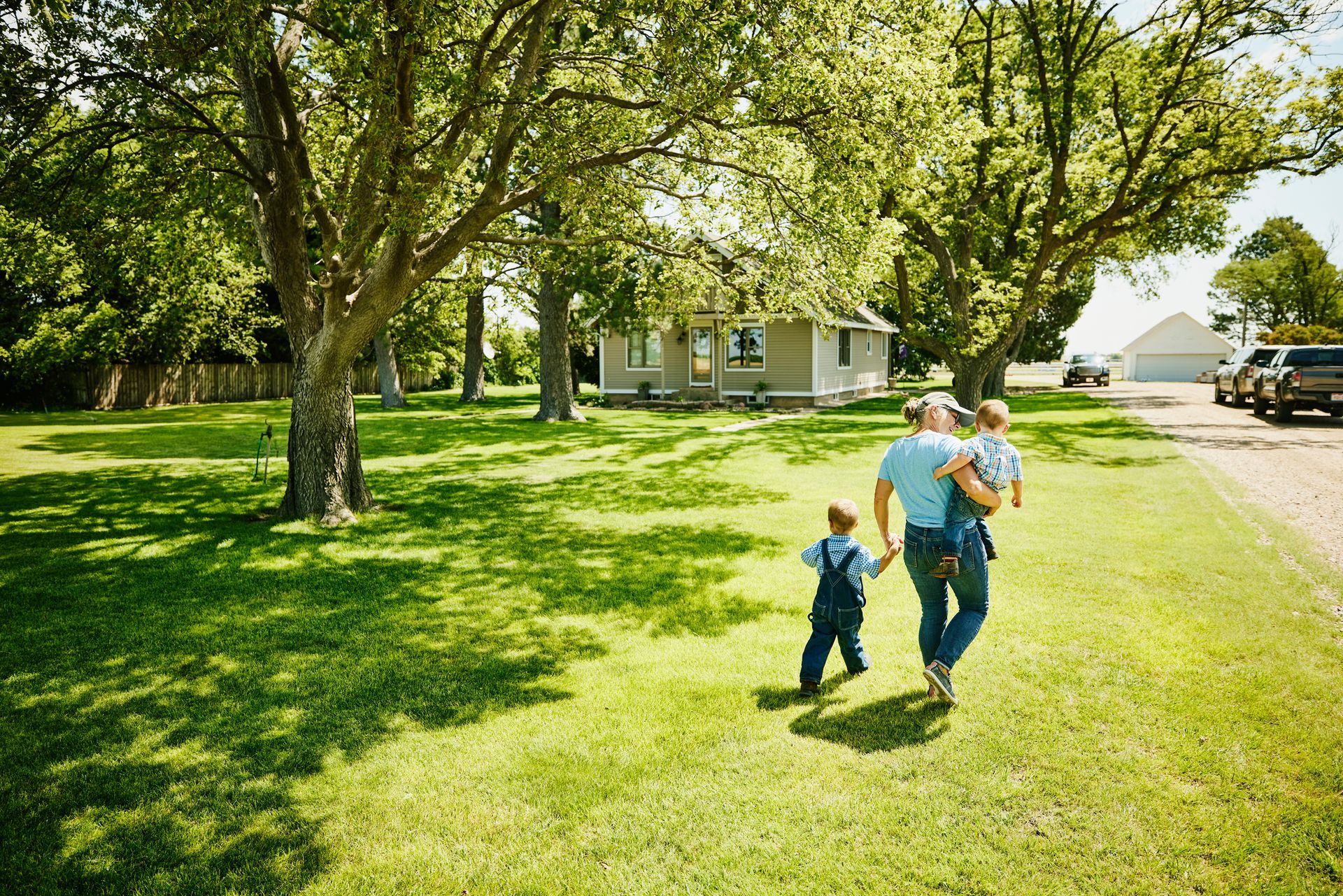 A woman walks with two children on a grassy lawn toward a house surrounded by trees on a sunny day.