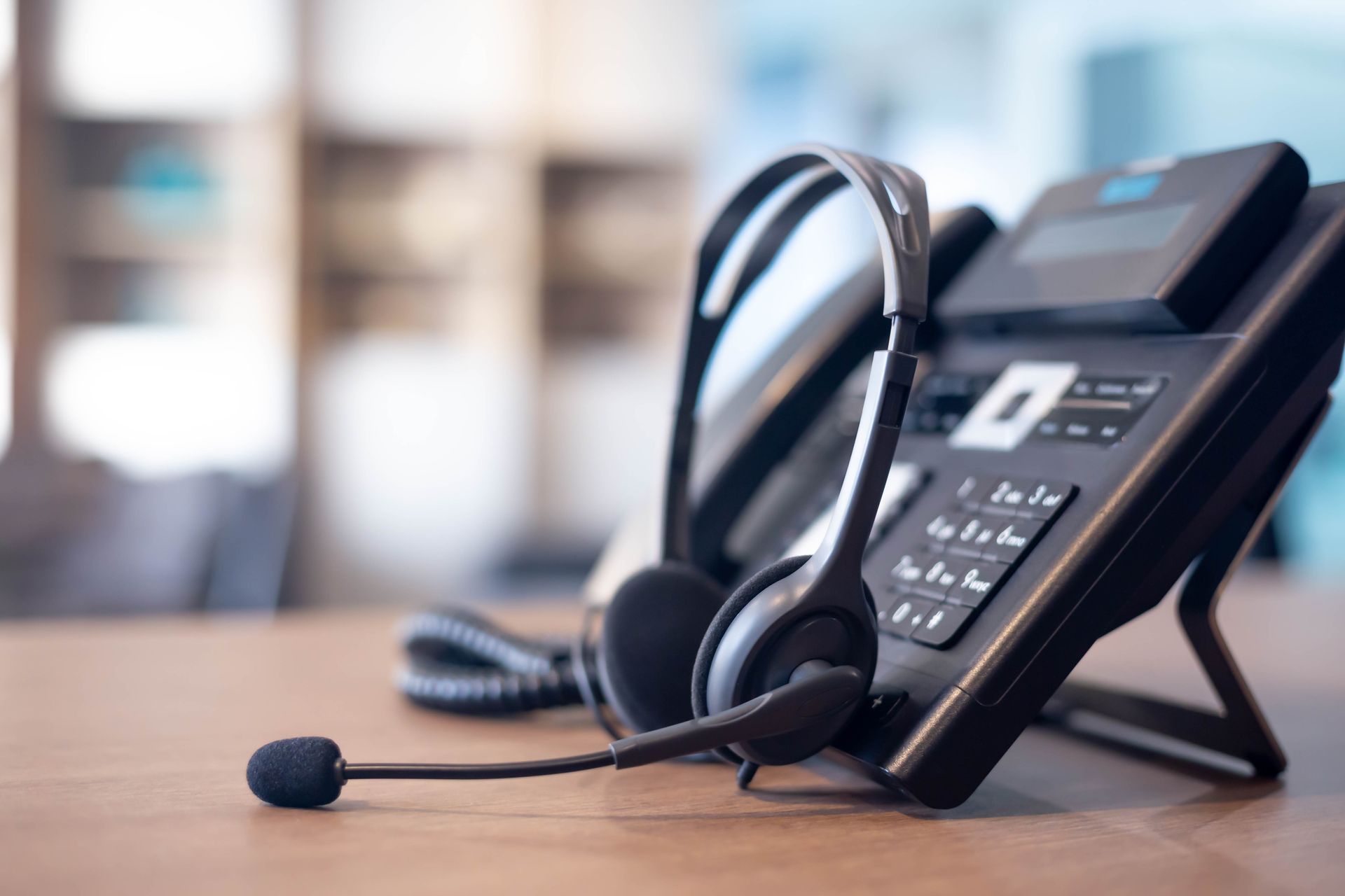 A black professional headset rests on a desk in front of a modern office desk phone.
