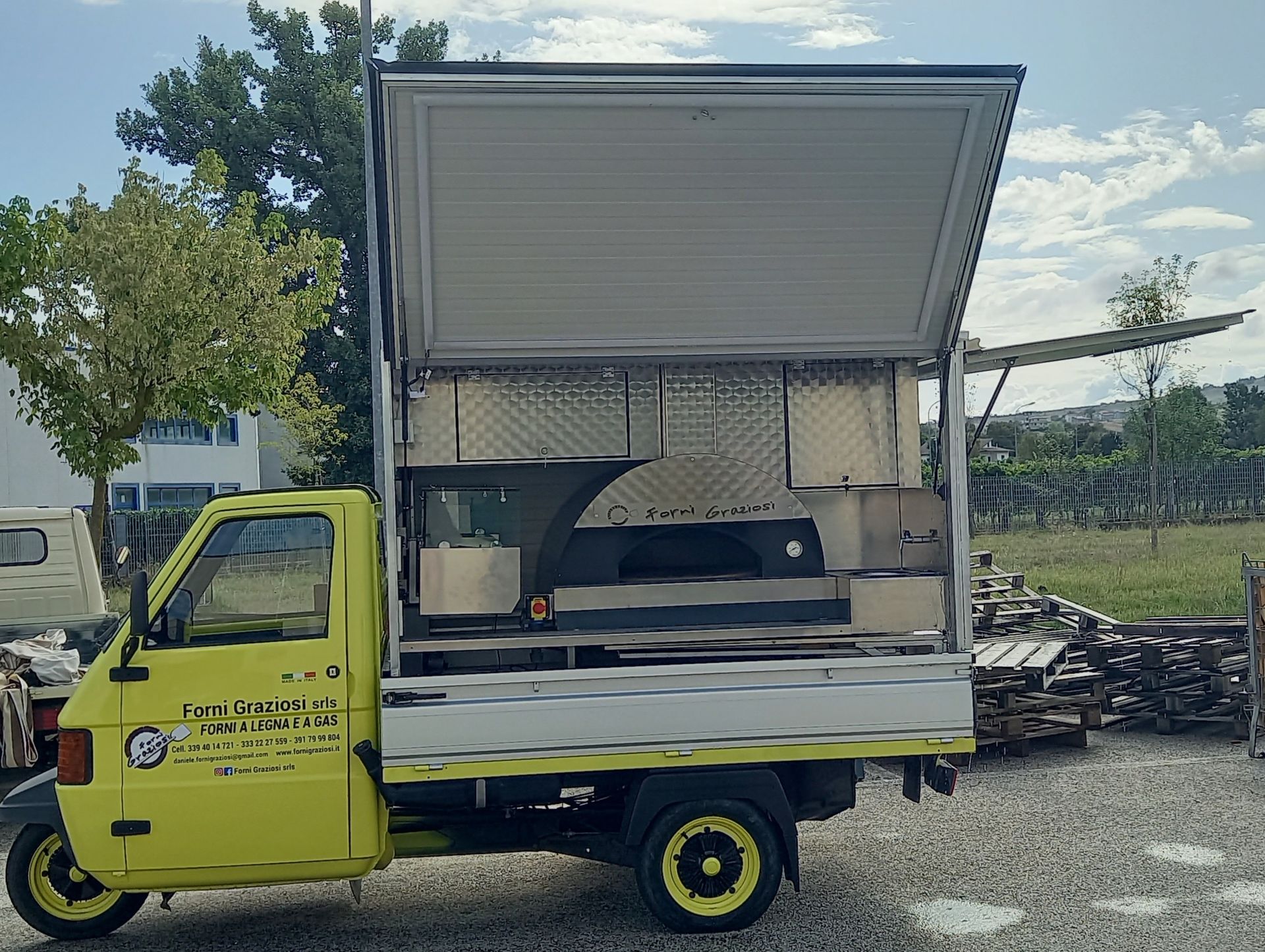 Un camion giallo con un forno per pizza sul retro è parcheggiato in uno spiazzo ghiaioso.