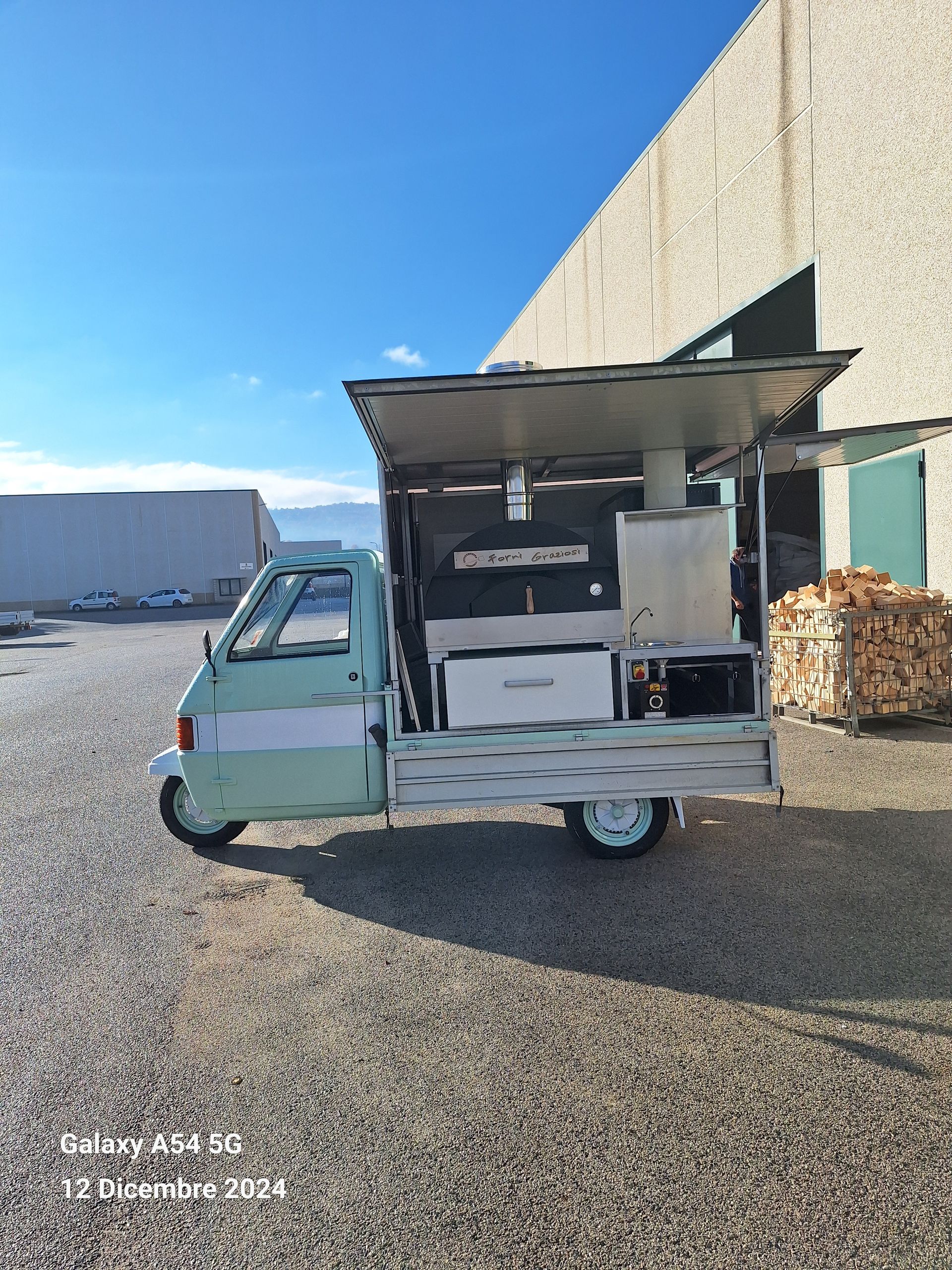 Un camioncino che vende cibo verde è parcheggiato in uno spiazzo ghiaioso di fronte a un edificio.
