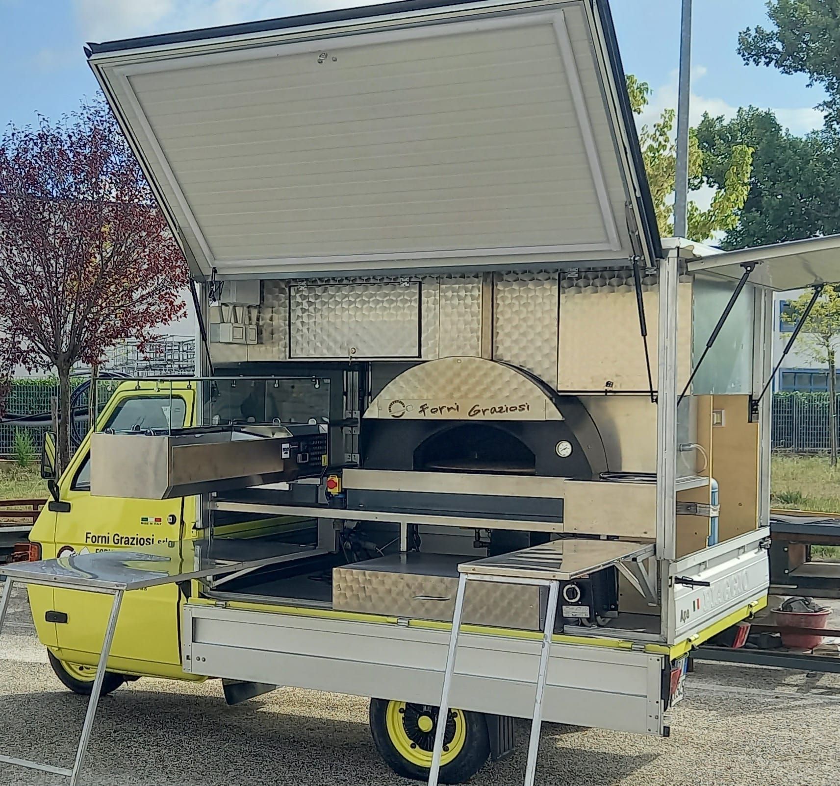Un camioncino giallo che vende cibo con un forno per pizza sul retro è parcheggiato in uno spiazzo ghiaioso.