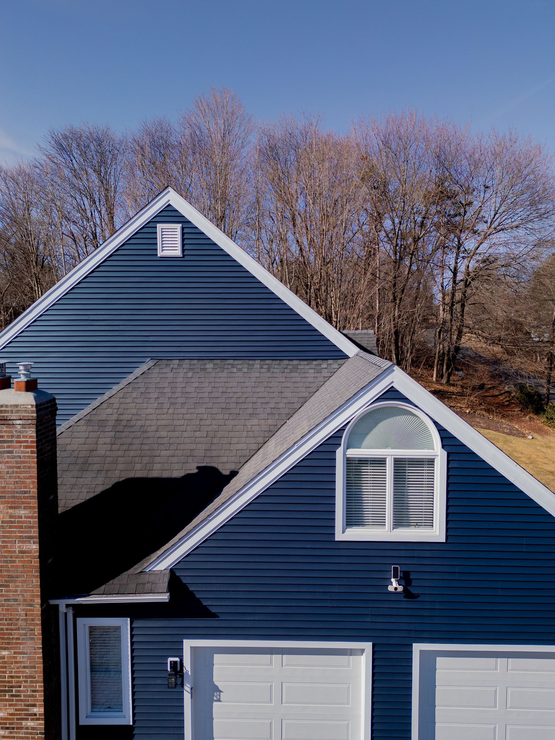 Blue-sided house with white garage doors and window, topped with a dark roof. Bare trees in the background, blue sky.
