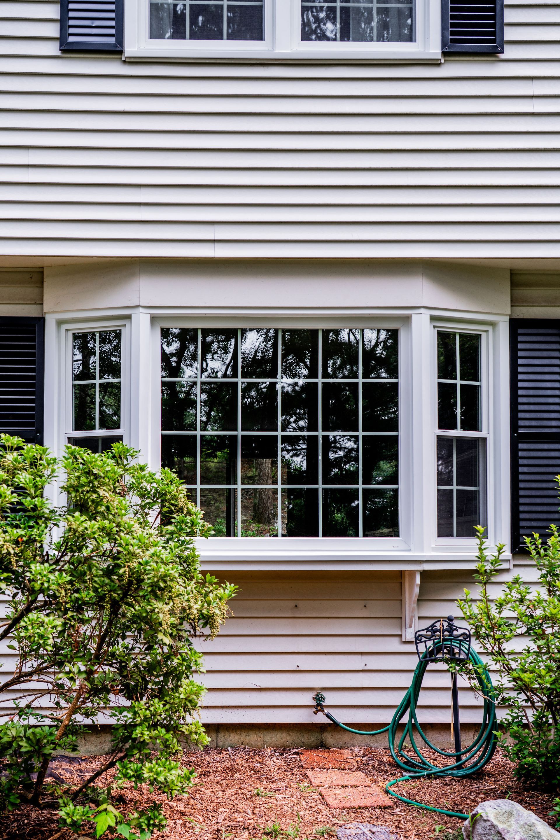 Bay window on a beige house with black shutters and green bushes.