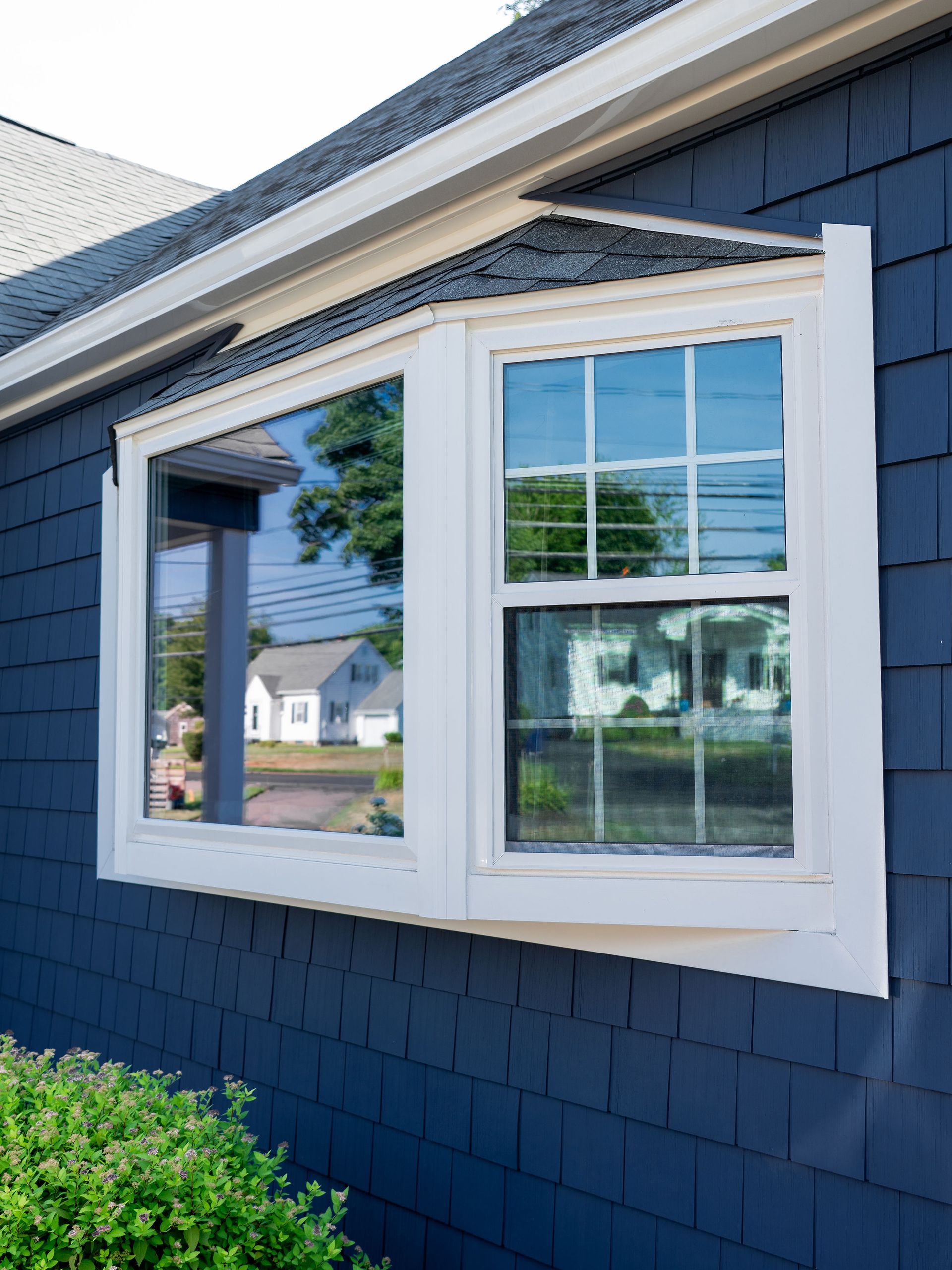 White-framed bay window on a blue shingled house; reflections of houses and trees visible.