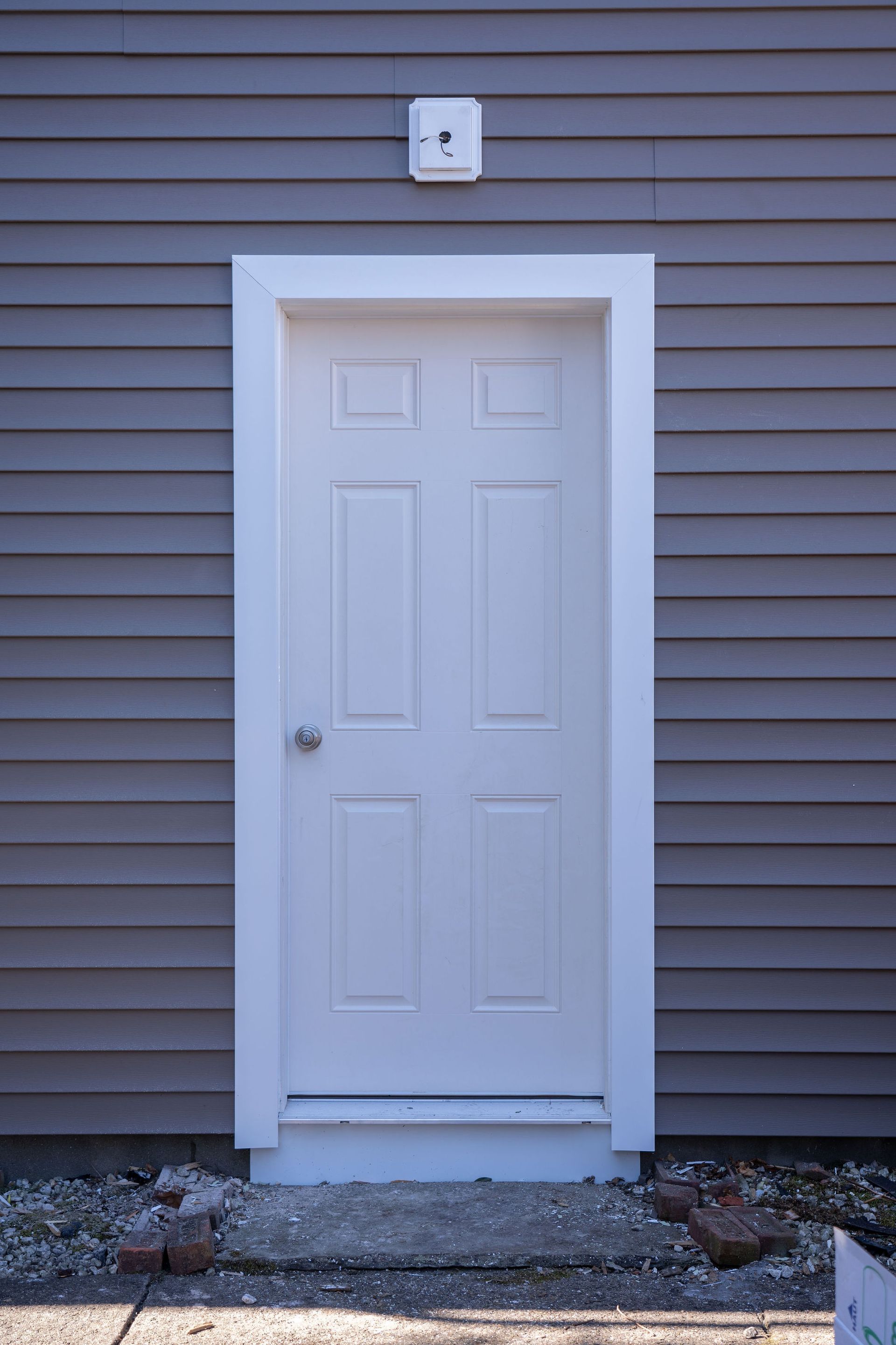 White six-panel door with white trim on gray siding, security camera above. Gravel and bricks at base.