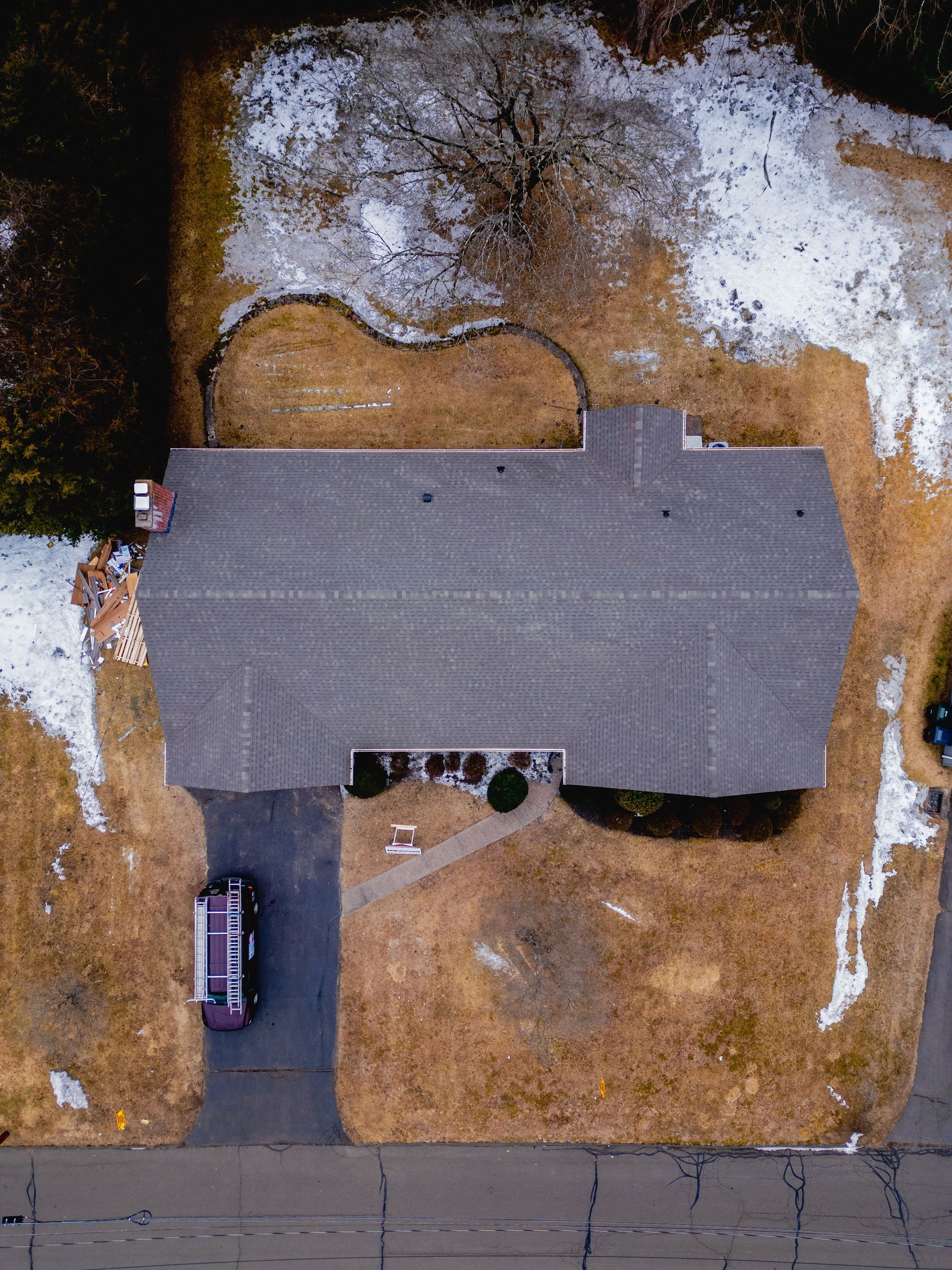 Aerial view of a house and yard with snow, brown grass, and a car parked in the driveway.
