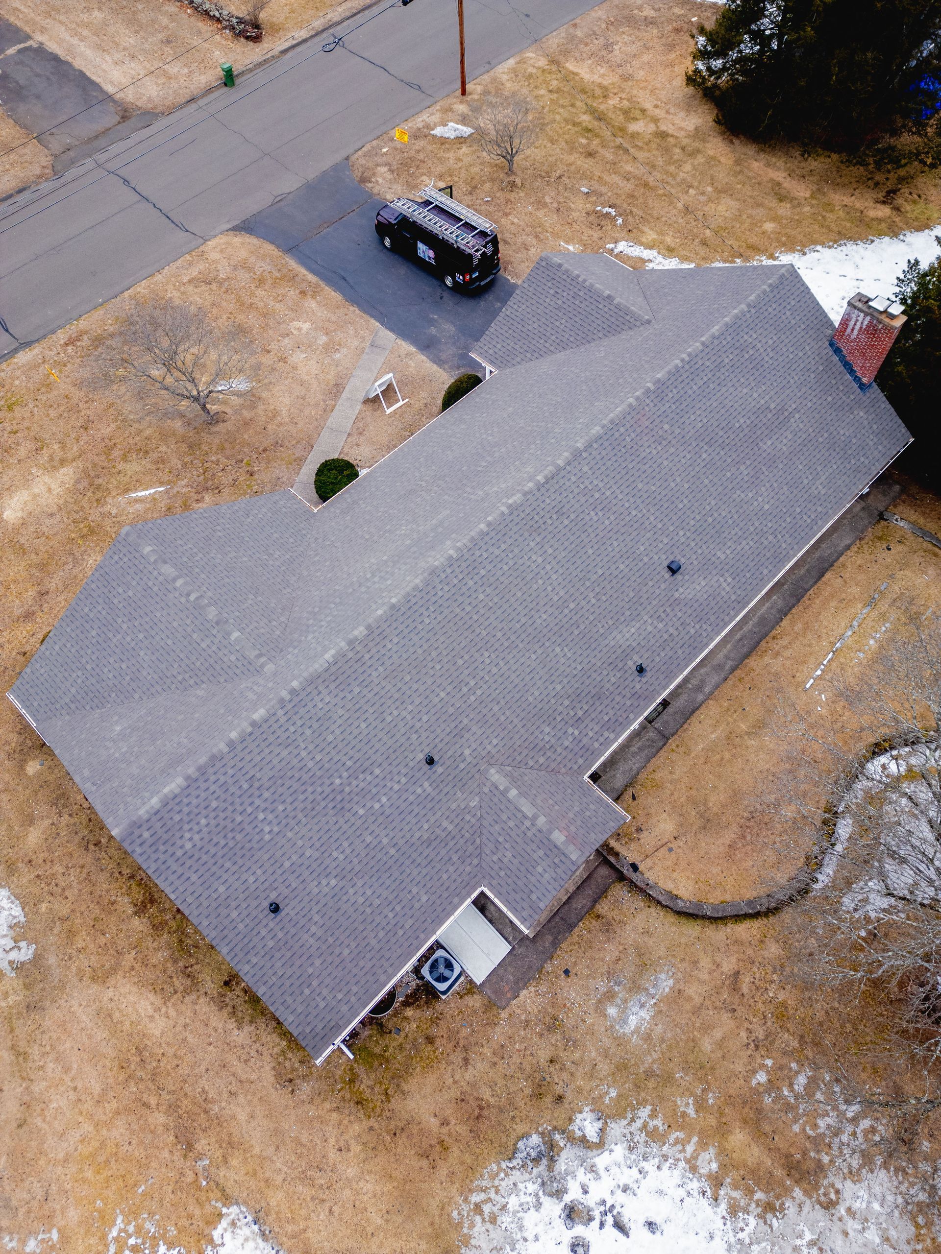 Aerial view of a house with a gray roof, driveway, and black car. Brown field surrounds the house.