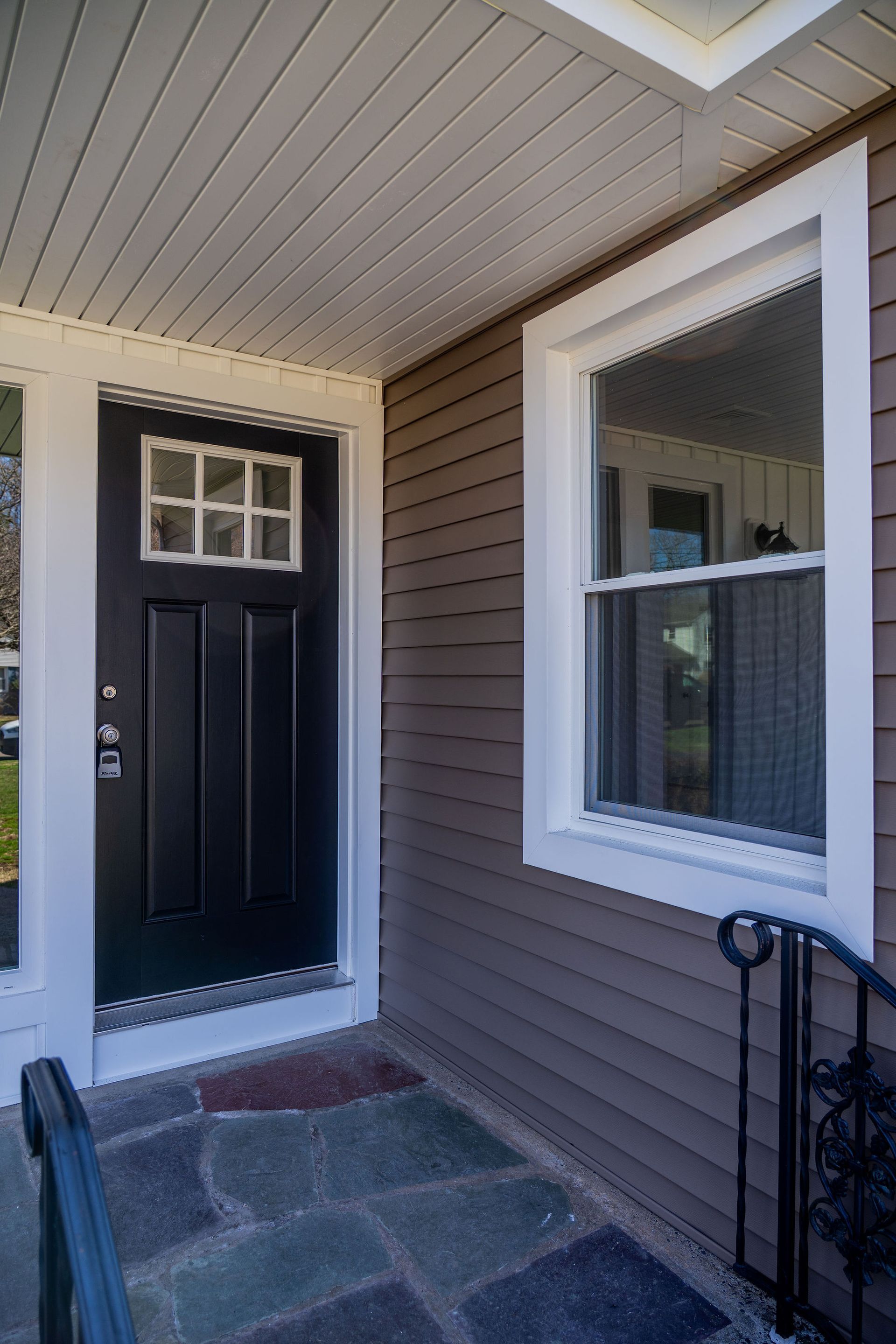Black front door with glass panes, next to a white-framed window and brown siding. Stone porch.