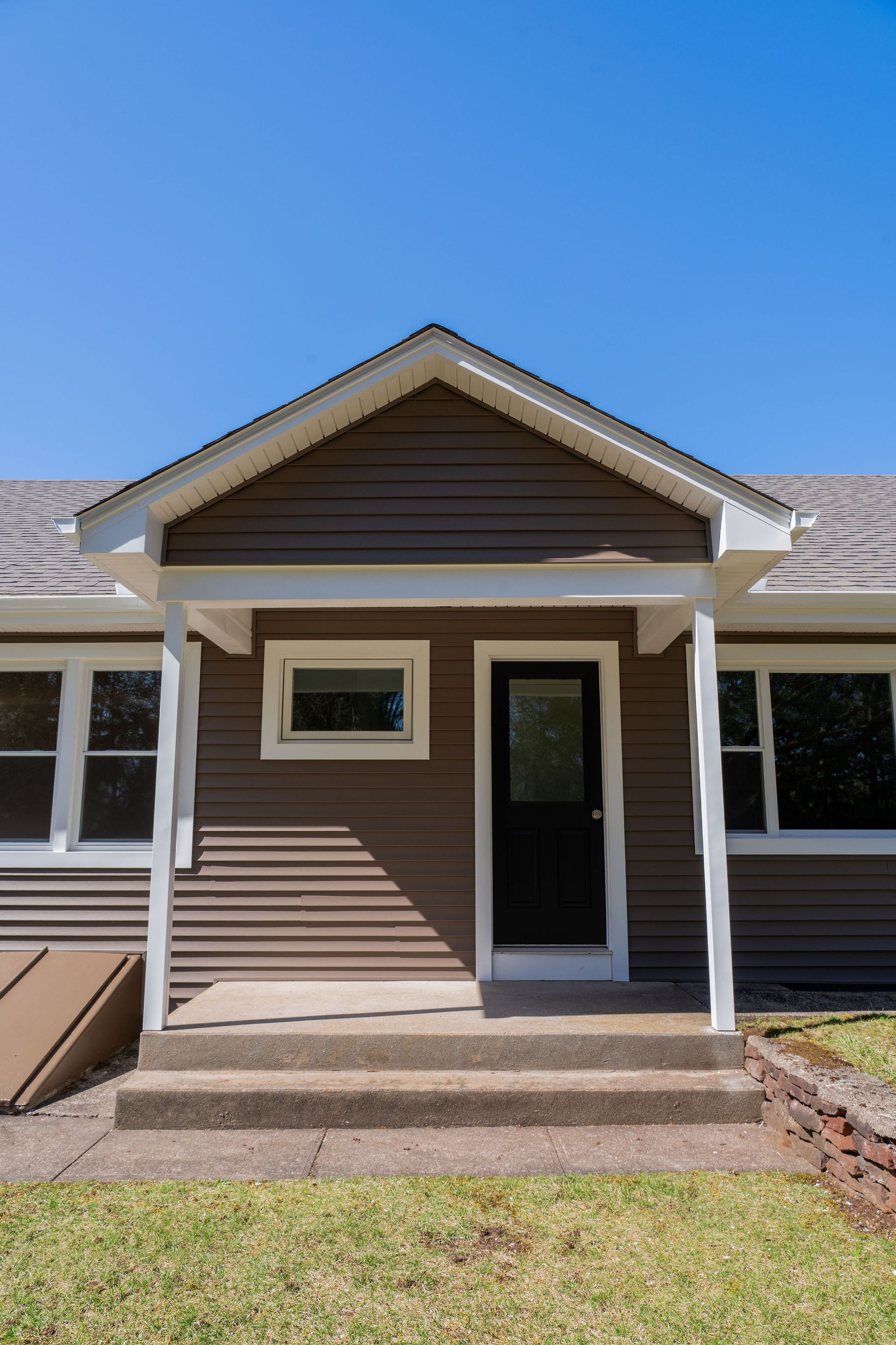 Brown house front with steps, black door, and white trim under a clear blue sky.